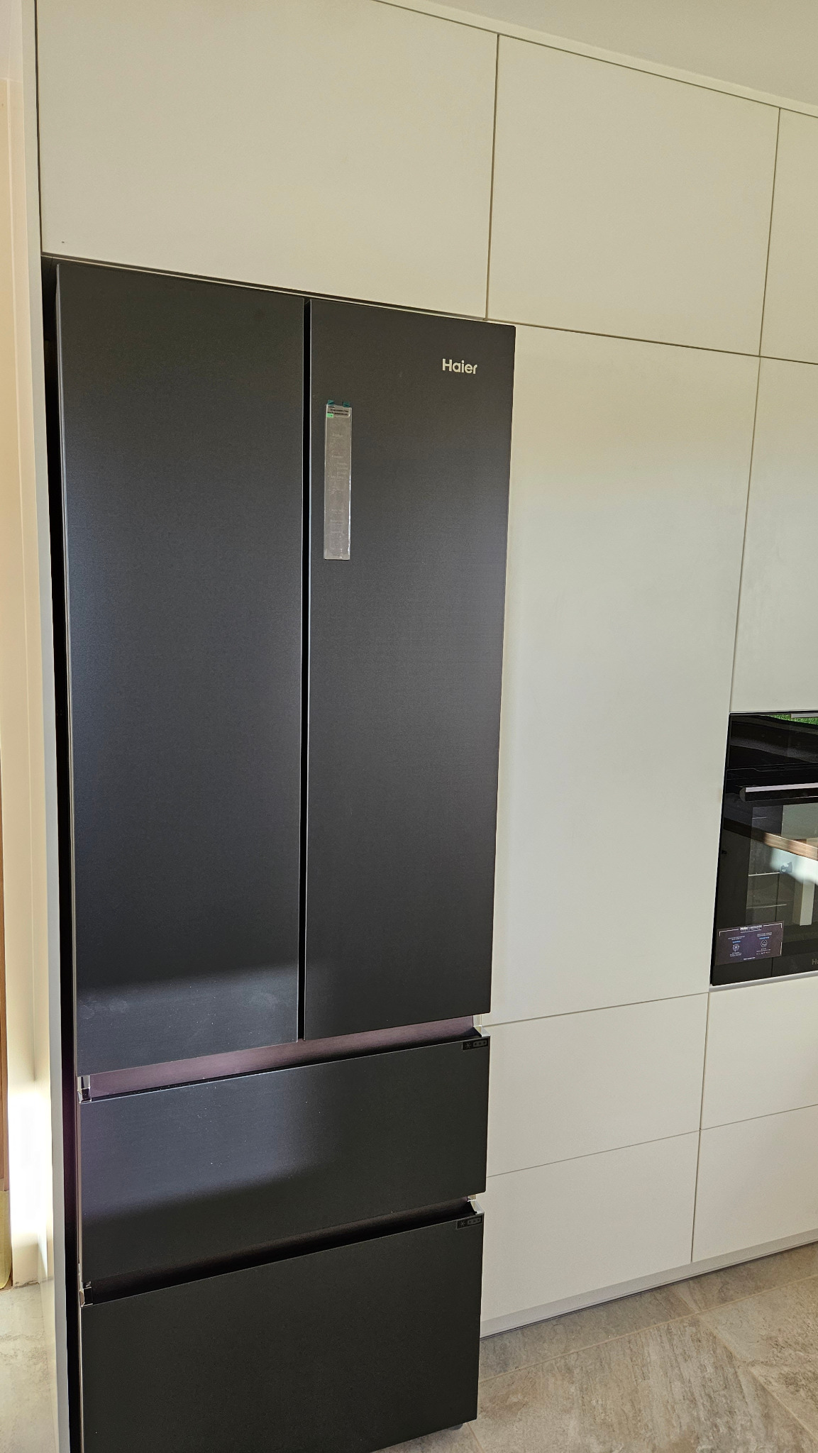 Czarna, nowoczesna lodówka Haier side-by-side w zabudowie kuchennej z białymi, matowymi frontami szafek.