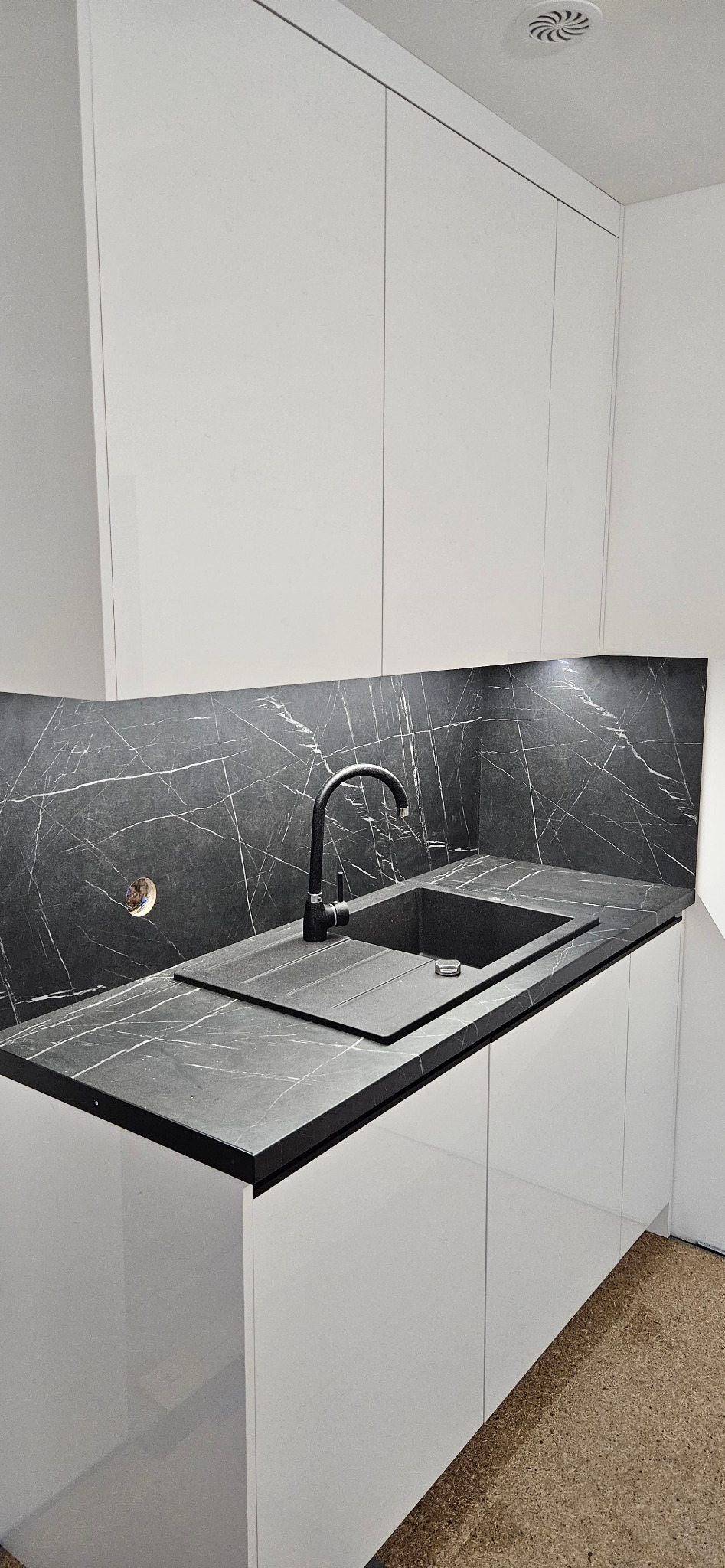 Minimalistyczna kuchnia: białe, gładkie szafki z czarnym blatem imitującym marmur i czarnym zlewem z baterią.