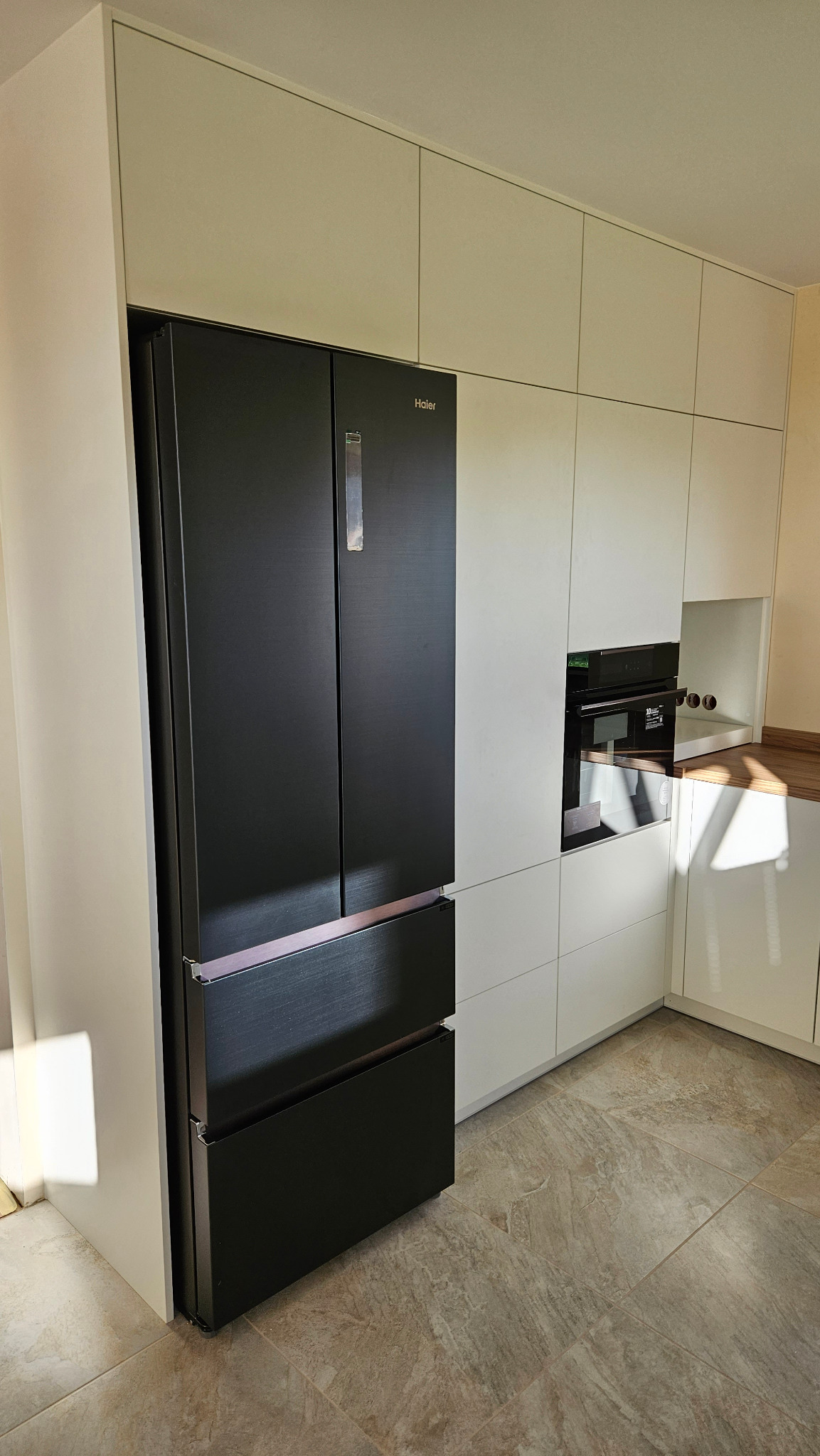 Czarna, czterodrzwiowa lodówka Haier w zabudowie z białych szafek kuchennych z piekarnikiem i drewnianym blatem, widok z boku na tle płytek podłogowych imitujących kamień.