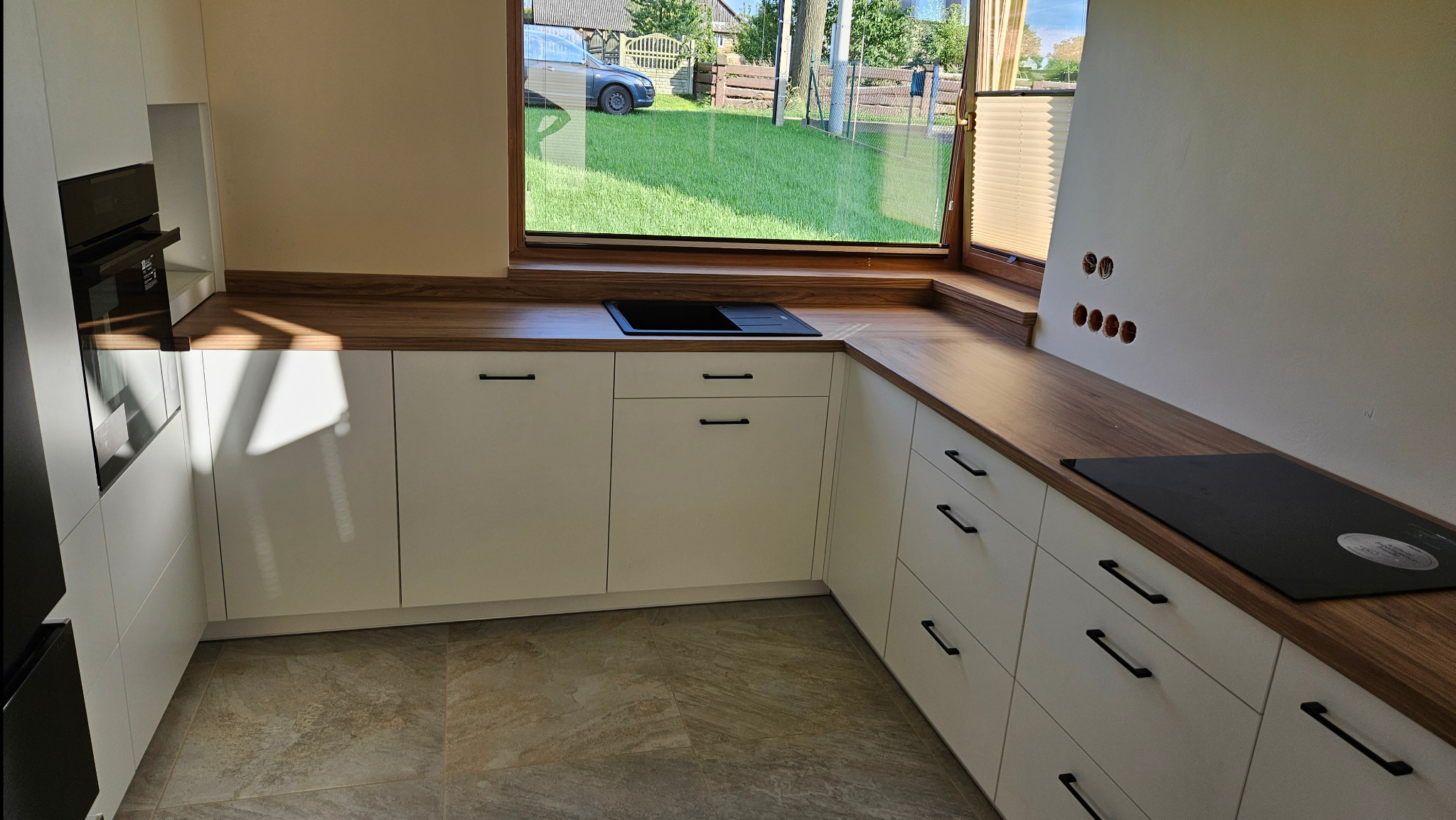 Nowoczesna zabudowa kuchenna z białymi frontami i drewnianym blatem, czarny zlew wpuszczany pod oknem z widokiem na ogród.