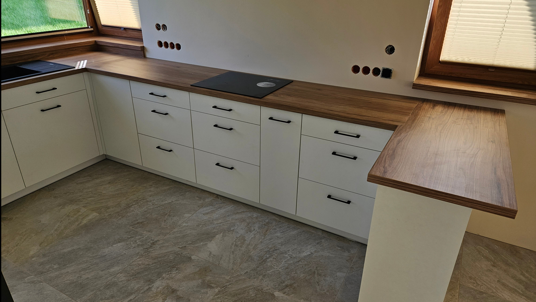 Białe szafki kuchenne z drewnianym blatem, czarne uchwyty, płyta indukcyjna, okno ze żaluzjami, widoczne gniazdka elektryczne w ścianie.