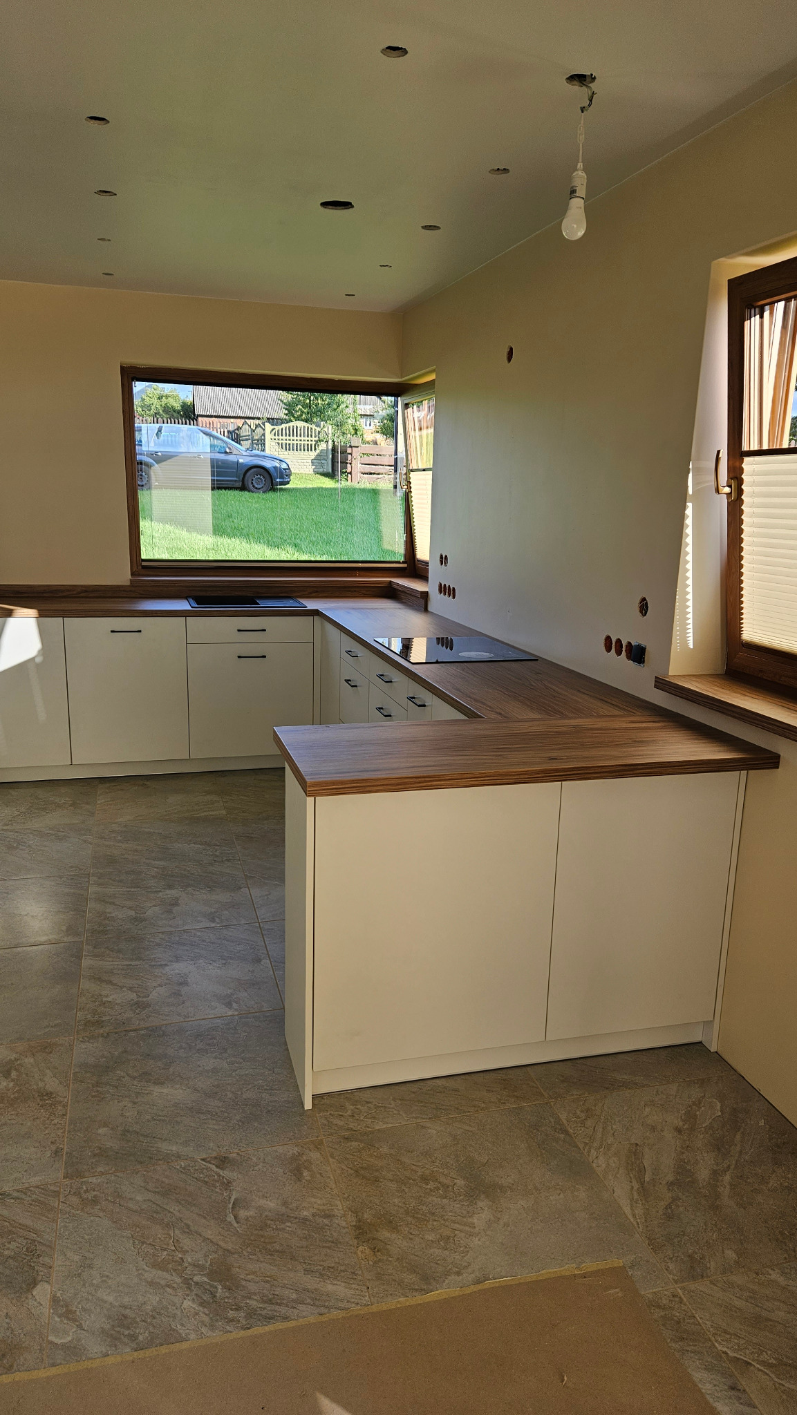 Wnętrze kuchni w trakcie wykańczania, z białymi szafkami i drewnianym blatem, widoczne przygotowane otwory na gniazdka elektryczne i wisząca żarówka.