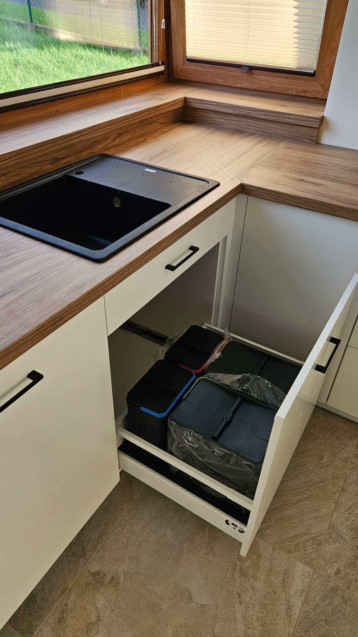 Wysuwana szuflada szafki pod zlewem z sortownikami na śmieci, widoczny blat z drewnopodobnym wykończeniem, czarny zlew i okno z widokiem na zieleń.