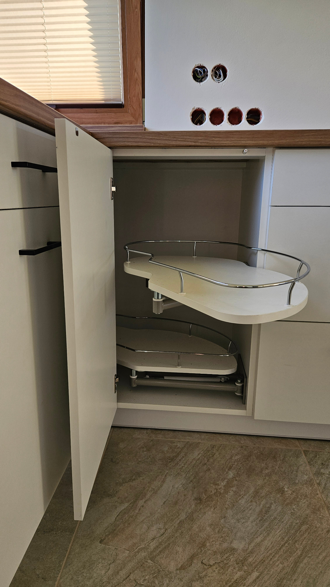 Wnętrze szafki narożnej z półkami obrotowymi, białe fronty, widoczne otwory instalacyjne w ścianie nad blatem.