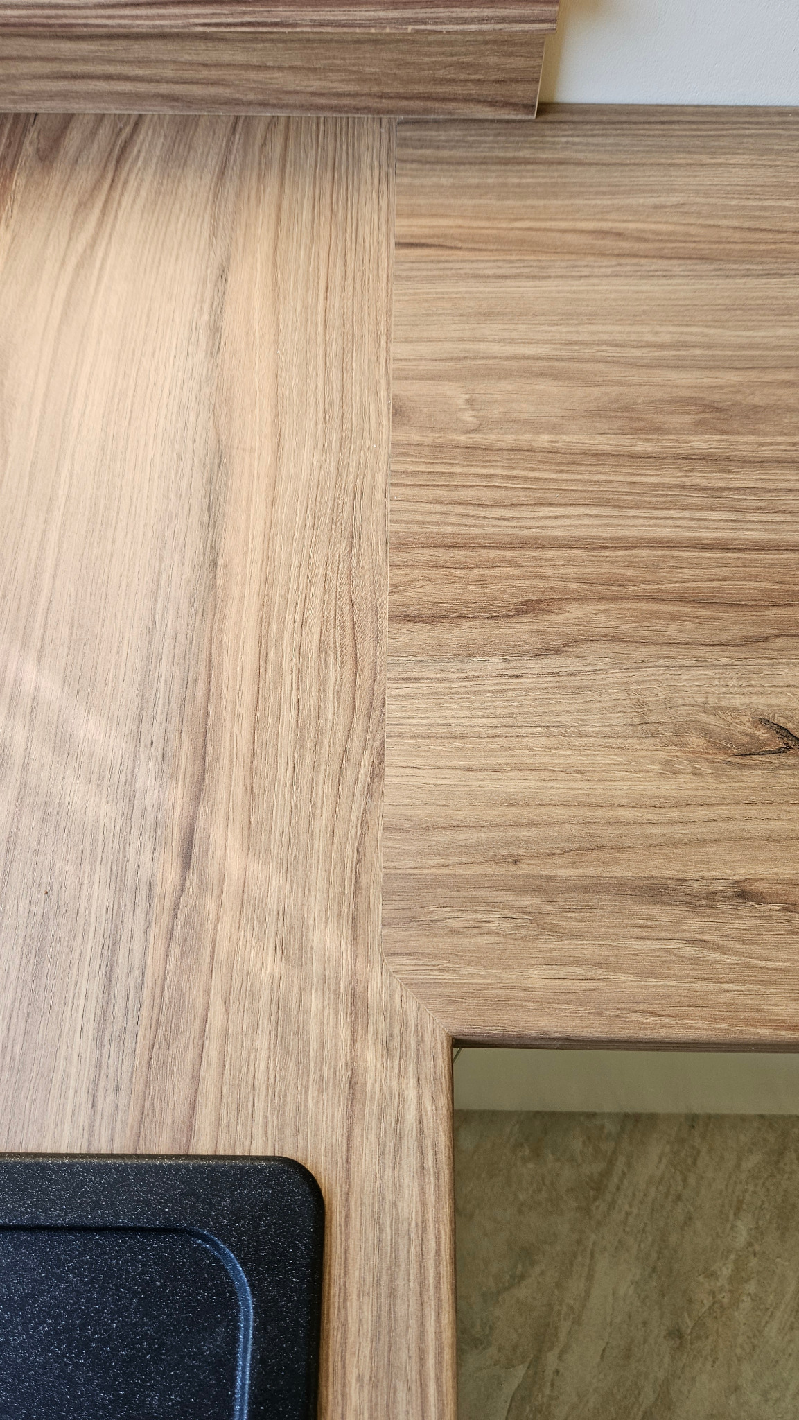 Szczegółowe zbliżenie na blat kuchenny z dwoma odcieniami drewna, z widocznym fragmentem zlewu w lewym dolnym rogu i fragmentem ściany w tle.