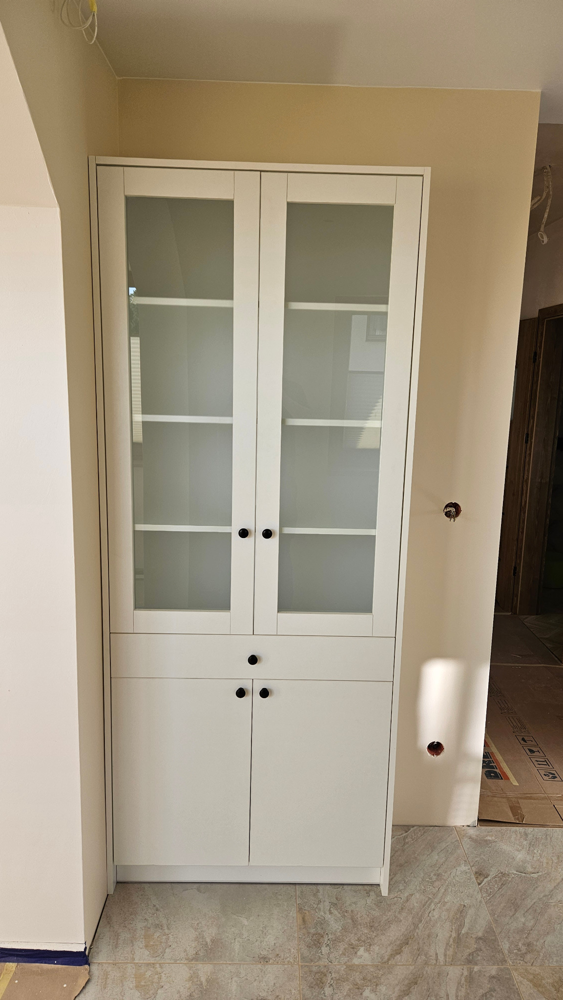 Biała przeszklona szafa z szufladami i szafkami w trakcie montażu, widoczne niedokończone wykończenie ścian i instalacja elektryczna.