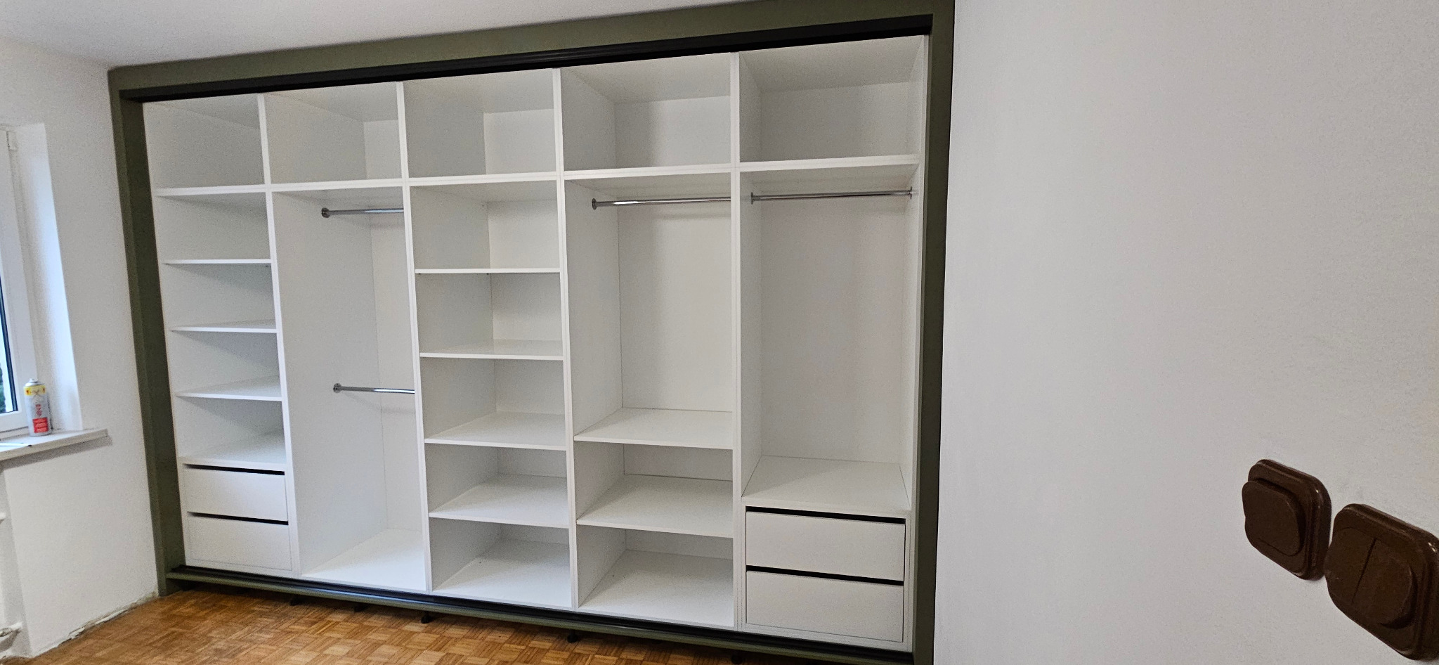 Wnętrze białej, otwartej szafy wnękowej z przesuwnymi drzwiami, widoczne półki, drążki i szuflady, rama w kolorze khaki, parkiet na podłodze.