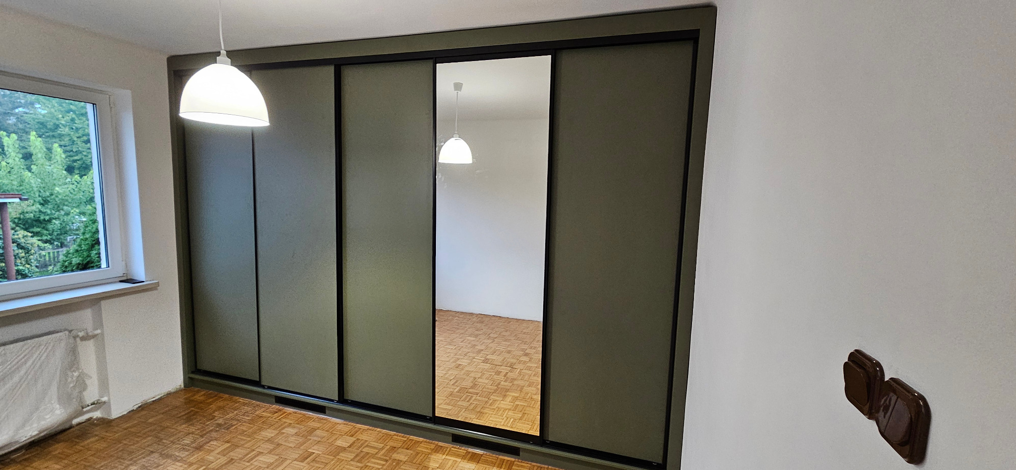 Zielona szafa przesuwna z lustrem w pokoju z parkietem, widoczne gniazdko elektryczne i lampa sufitowa.