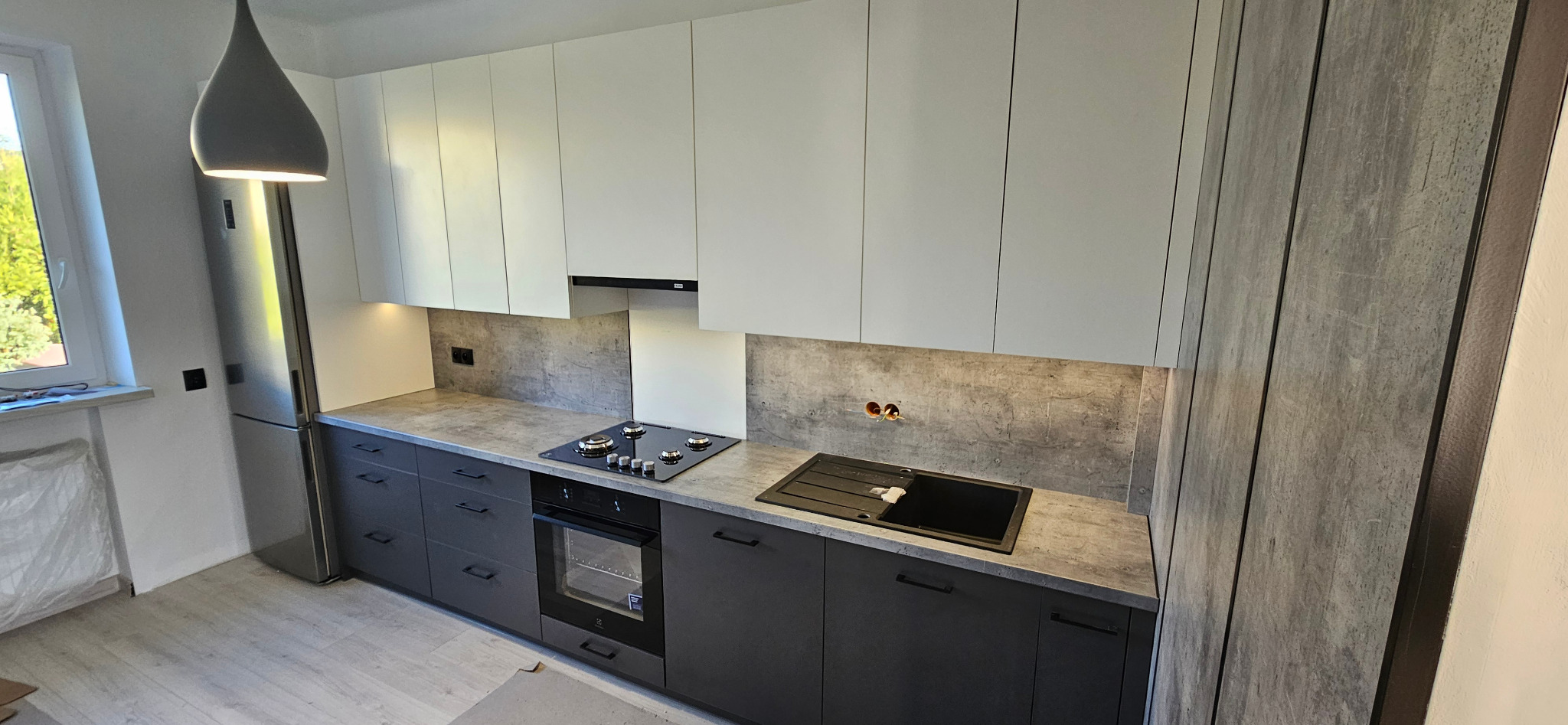 Nowoczesna kuchnia z białymi szafkami górnymi i grafitowymi dolnymi, szarym blatem i panelem ściennym imitującym beton, oświetlenie podszafkowe, wisząca lampa w kształcie kropli.