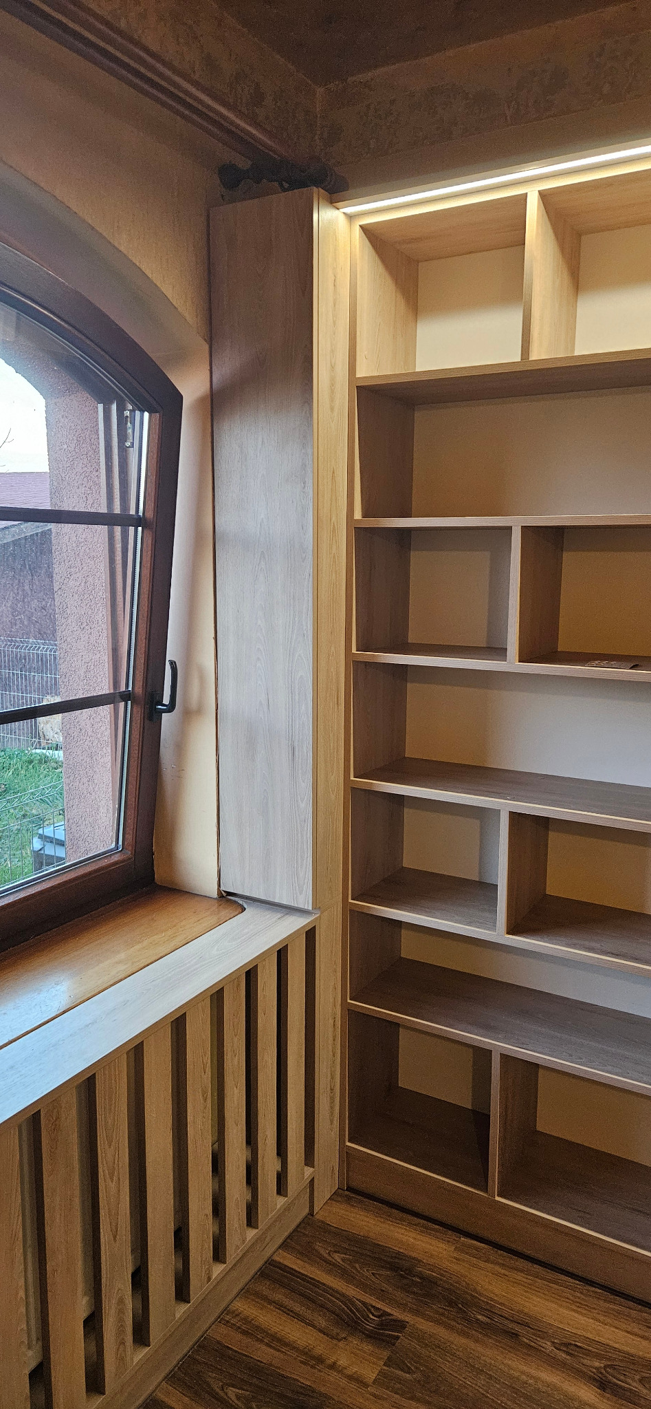 Jasna, drewniana zabudowa wnęki okiennej z grzejnikiem i regałem z oświetleniem LED w górnej części.