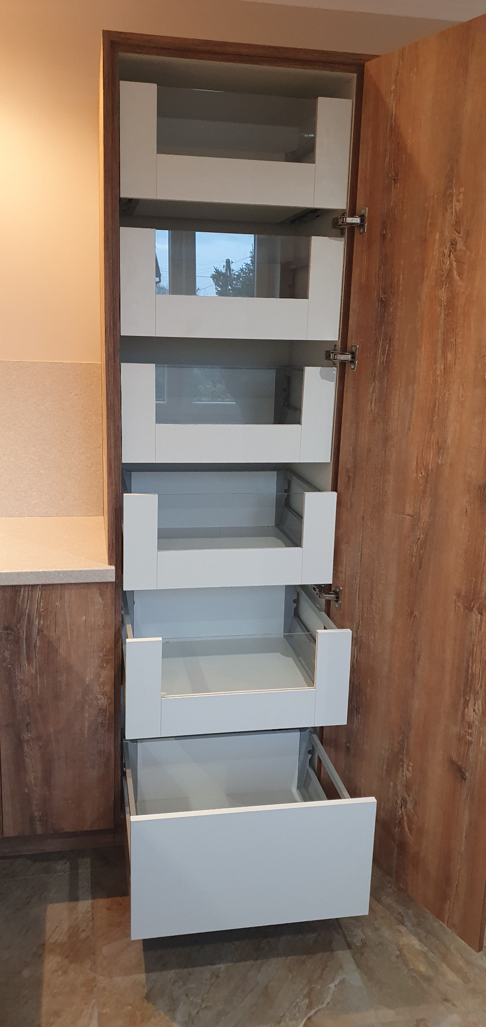 Wysoka szafka kuchenna z otwartymi, białymi szufladami z frontami z przezroczystego szkła, widoczne okucia, obok szafka z drewnopodobnym frontem.