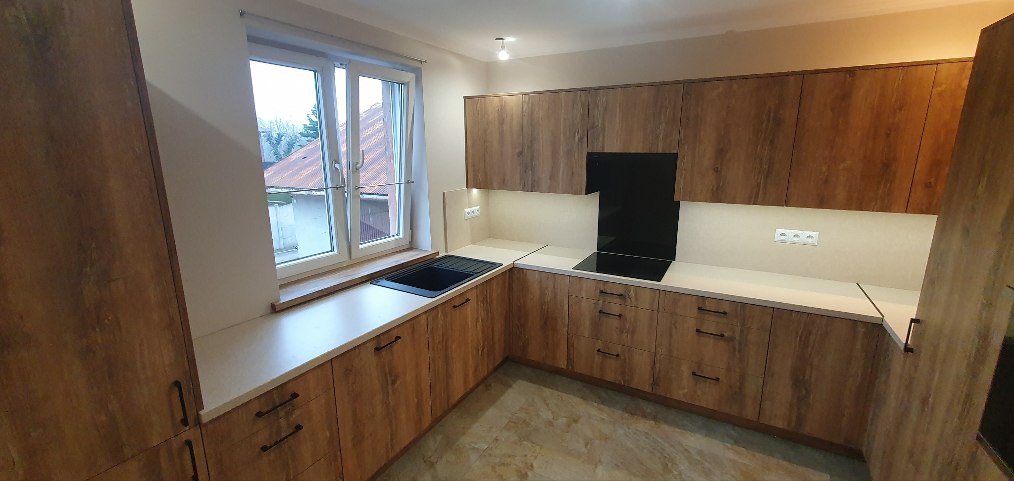 Nowoczesna kuchnia z drewnianymi frontami, czarnym zlewem pod oknem i płytą indukcyjną na białym blacie, oświetlenie podszafkowe.