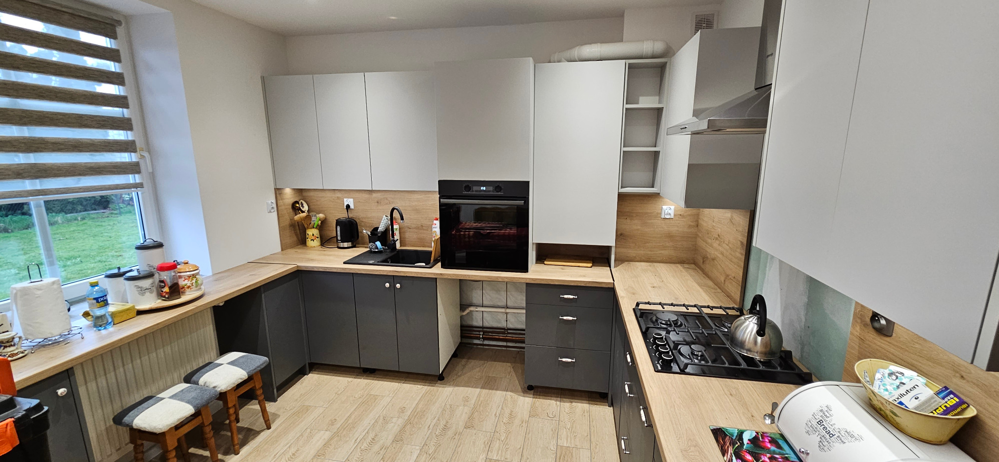 Nowoczesna kuchnia w odcieniach szarości i bieli z drewnianym blatem, widok na okno z roletami i ogród, czarny zlew i piekarnik, oraz kuchenka gazowa z czajnikiem.