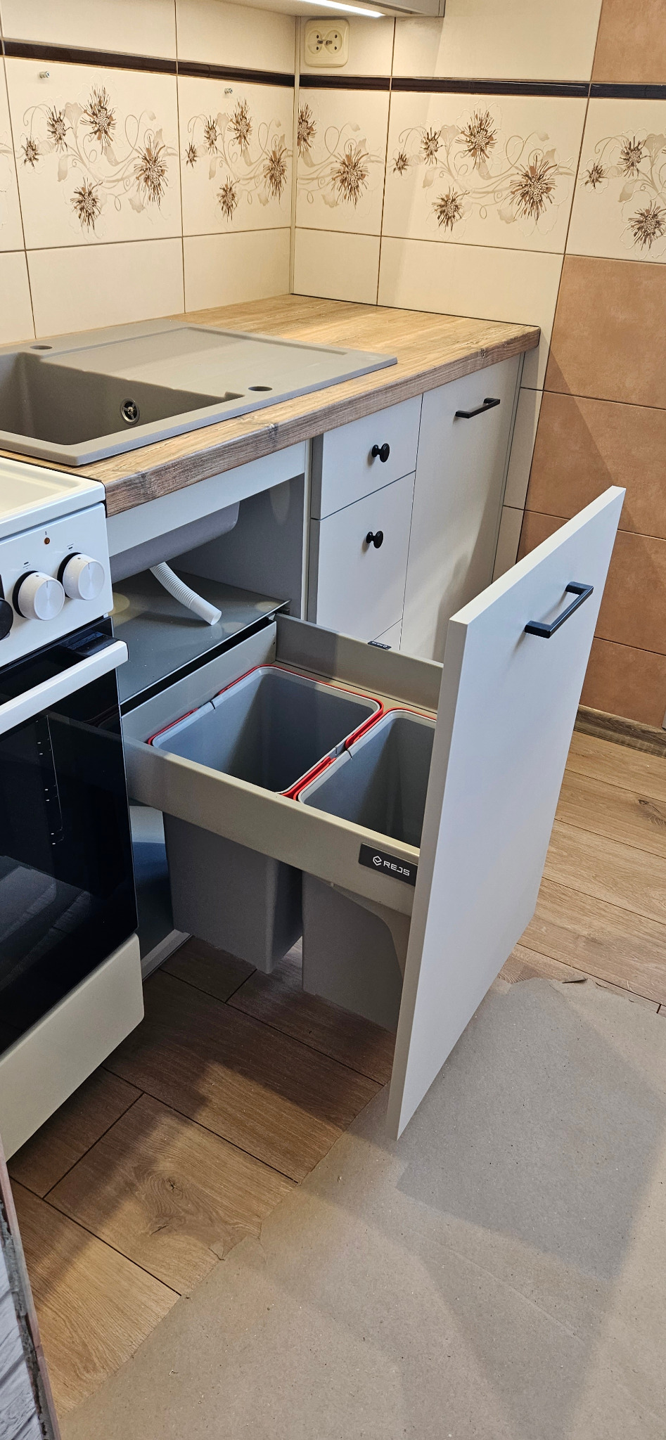 Szare szafki kuchenne z drewnianym blatem i wysuwanym koszem na śmieci firmy Rejs z dwoma pojemnikami, widoczne płytki ceramiczne z motywem kwiatowym