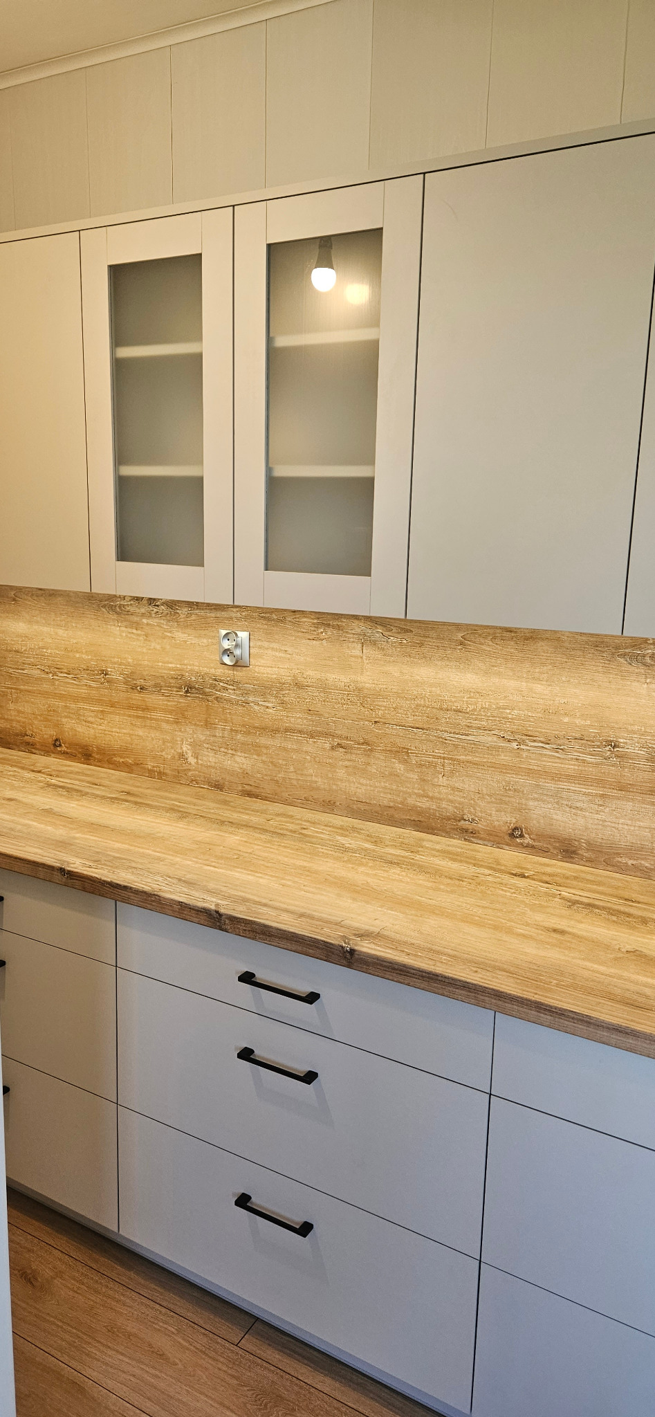 Jasnoszara zabudowa kuchenna z drewnianym blatem i gniazdkiem elektrycznym, widoczne przeszklone szafki górne i szuflady z czarnymi uchwytami.