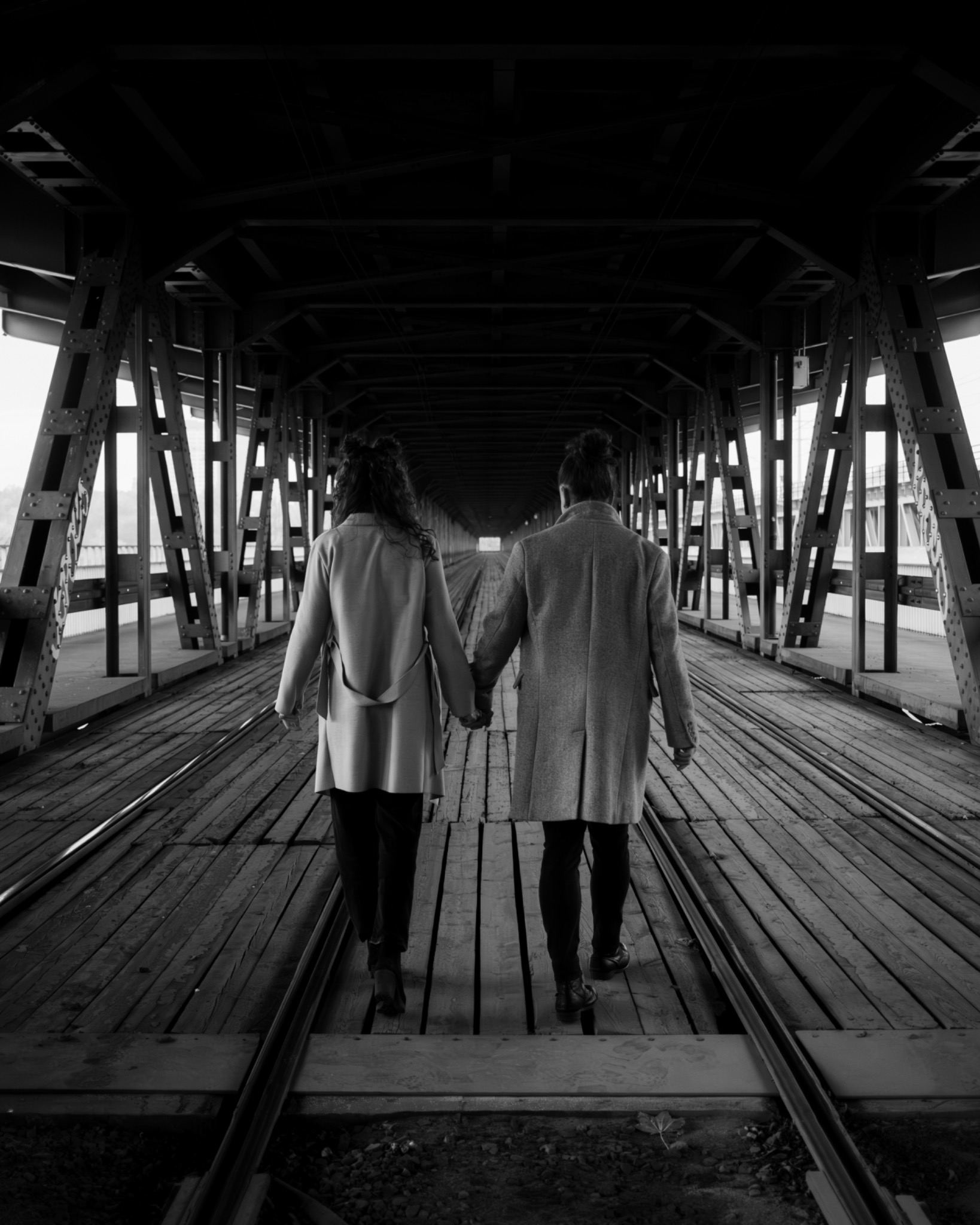 Para w płaszczach idzie trzymając się za ręce po drewnianym moście kolejowym, widok z tyłu, czarno-biała fotografia.