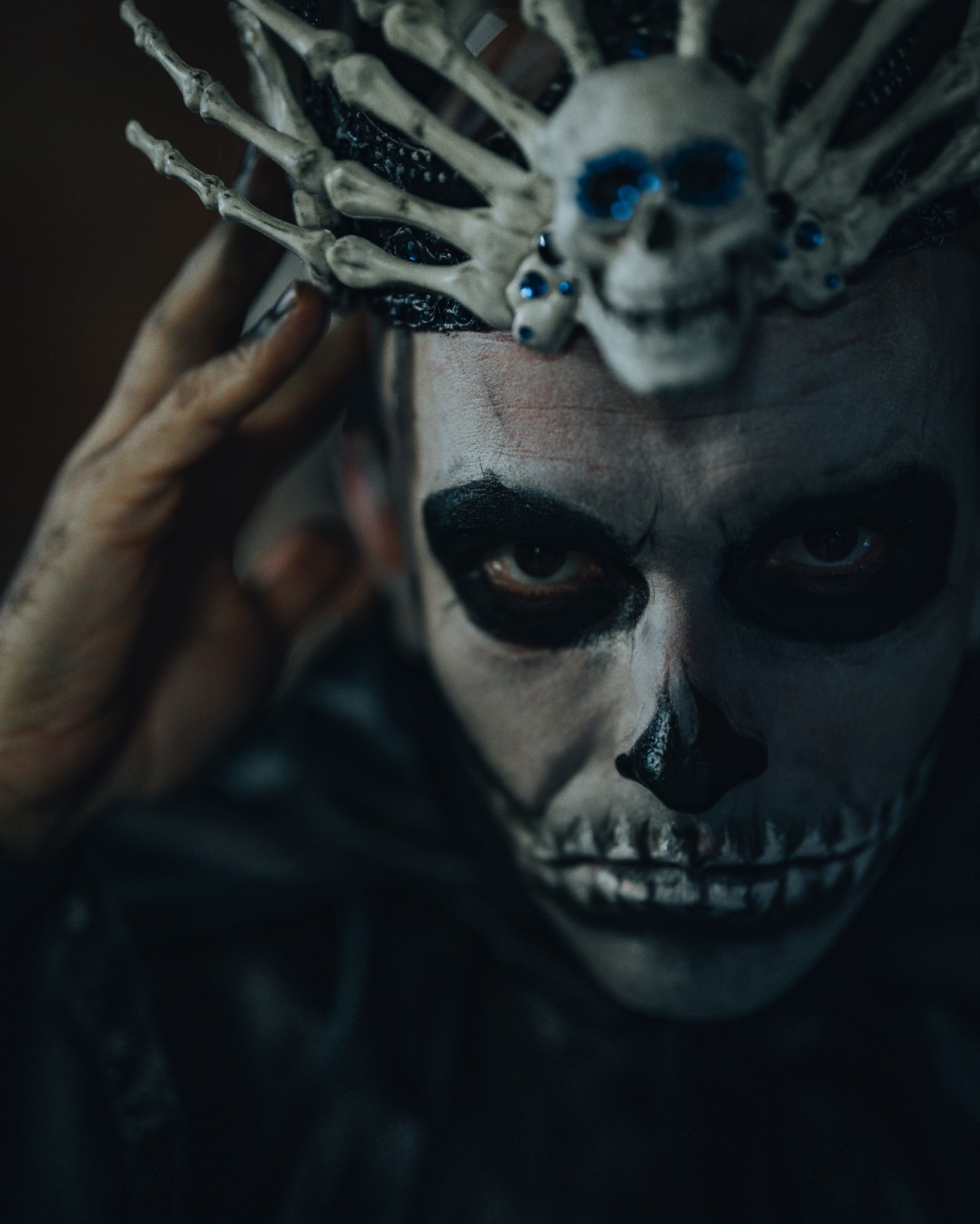 Portret osoby z makijażem czaszki i koroną z kości, z ręką uniesioną do głowy, w ciemnym oświetleniu.