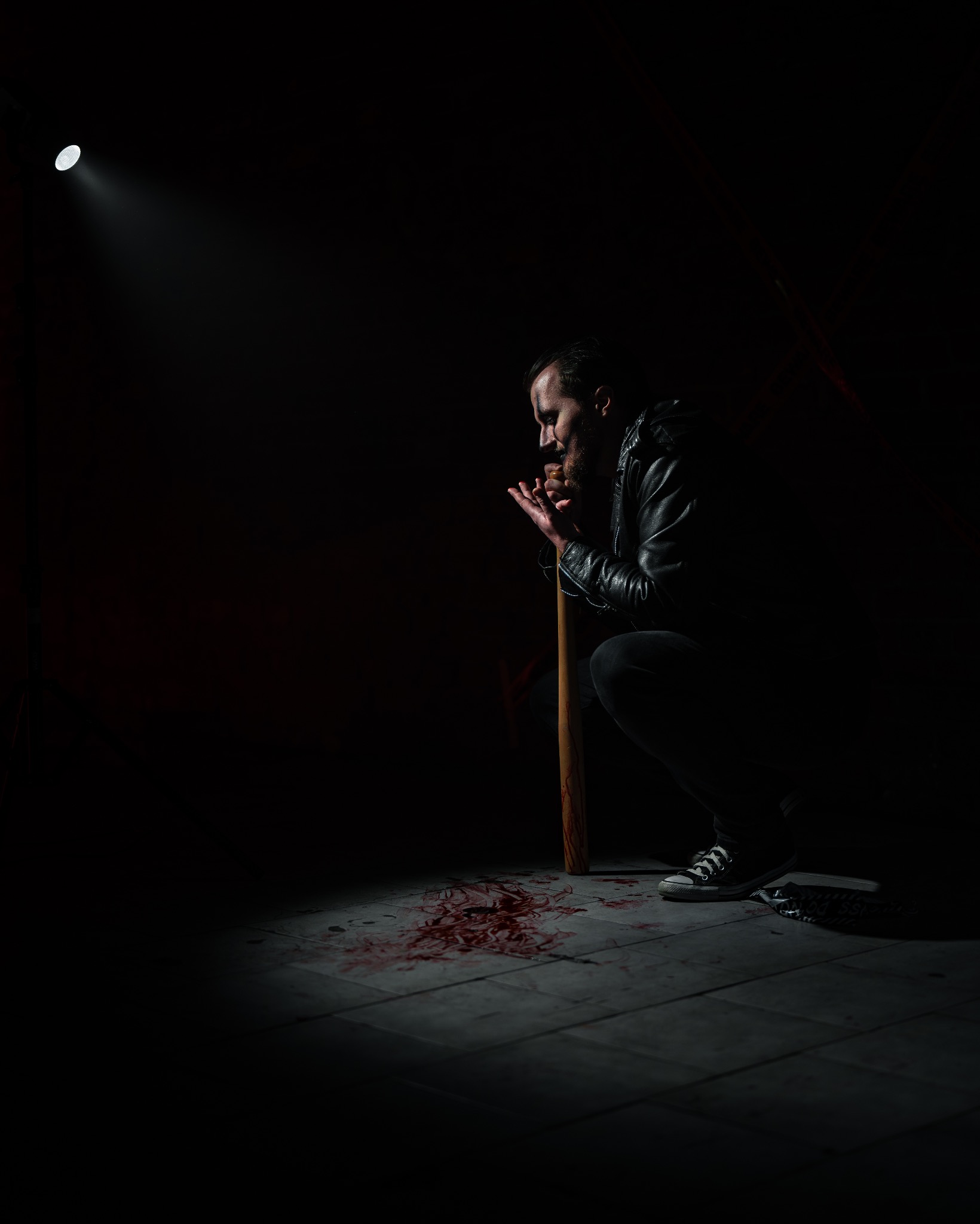 Mężczyzna w skórzanej kurtce kuca w ciemności, oświetlony punktowym światłem, trzymając w dłoniach kij bejsbolowy z plamami przypominającymi krew, podobne plamy na podłodze z płytek.