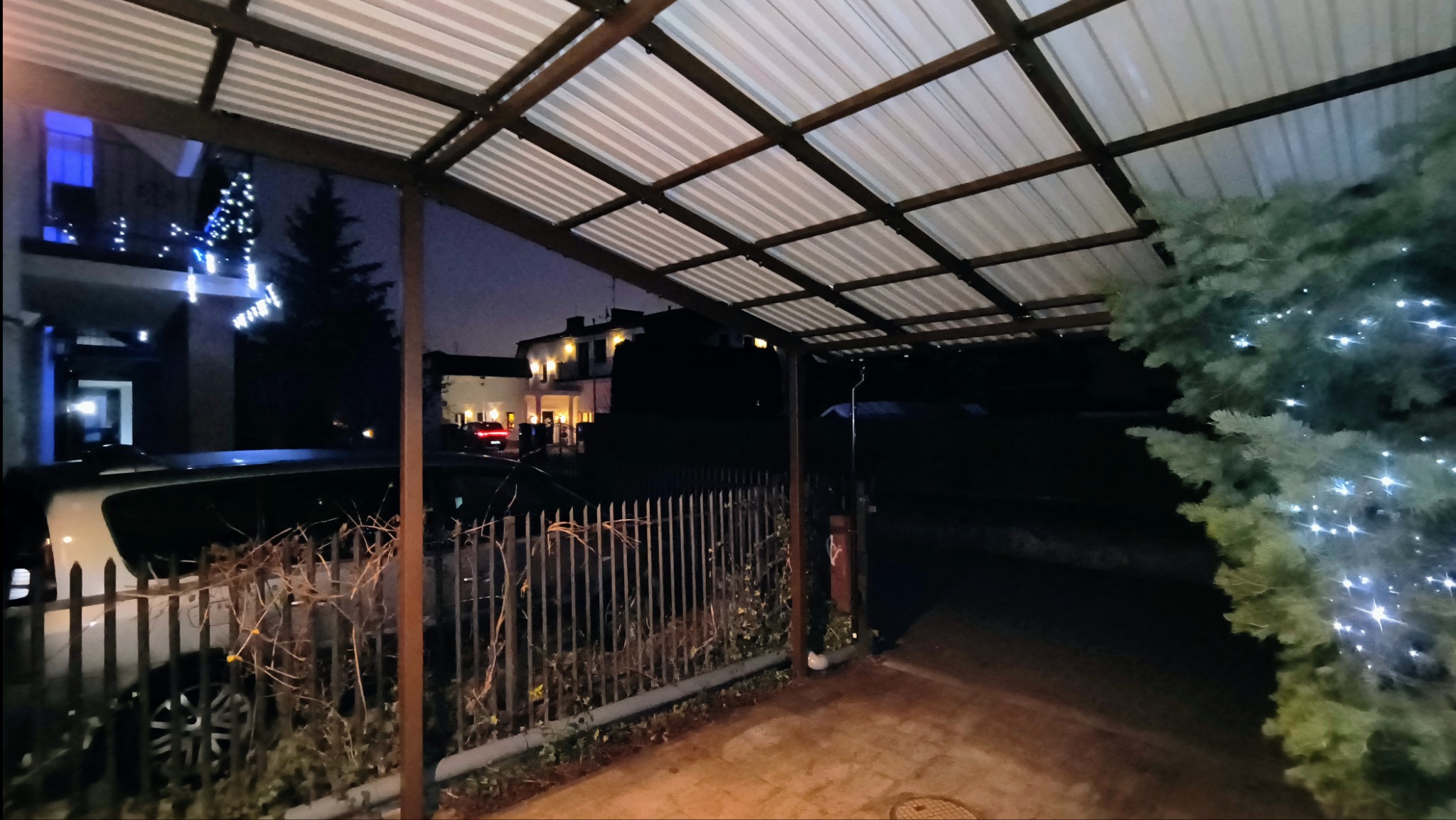 Metalowa konstrukcja zadaszenia z widocznymi elementami spawania, nocne ujęcie z samochodem za metalowym płotem i oświetlonym domem w tle, dekoracje świąteczne na budynku i drzewie.