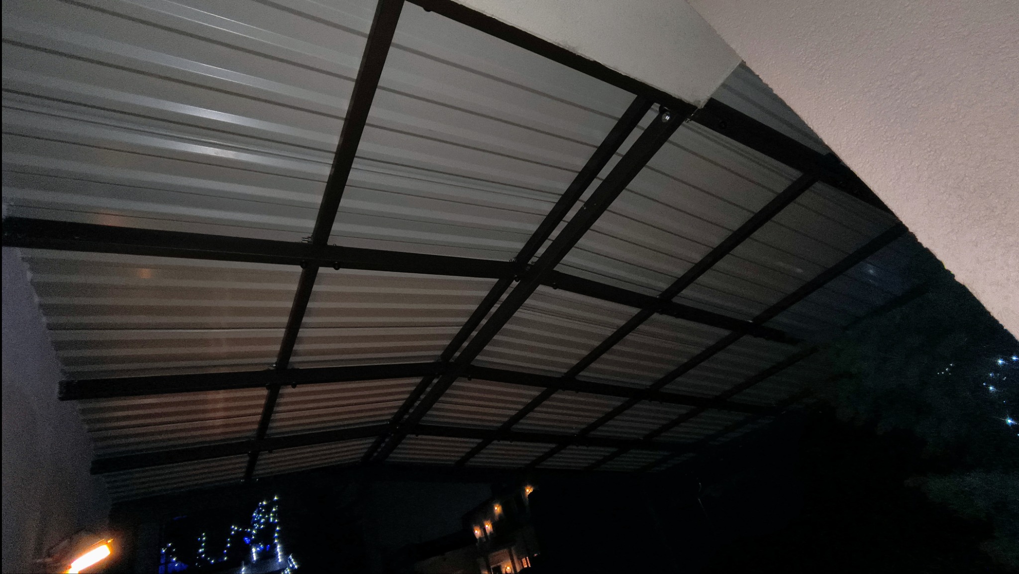 Metalowa konstrukcja z falistej blachy widziana od spodu, prawdopodobnie zadaszenie tarasu lub werandy, w tle nocne oświetlenie budynku.