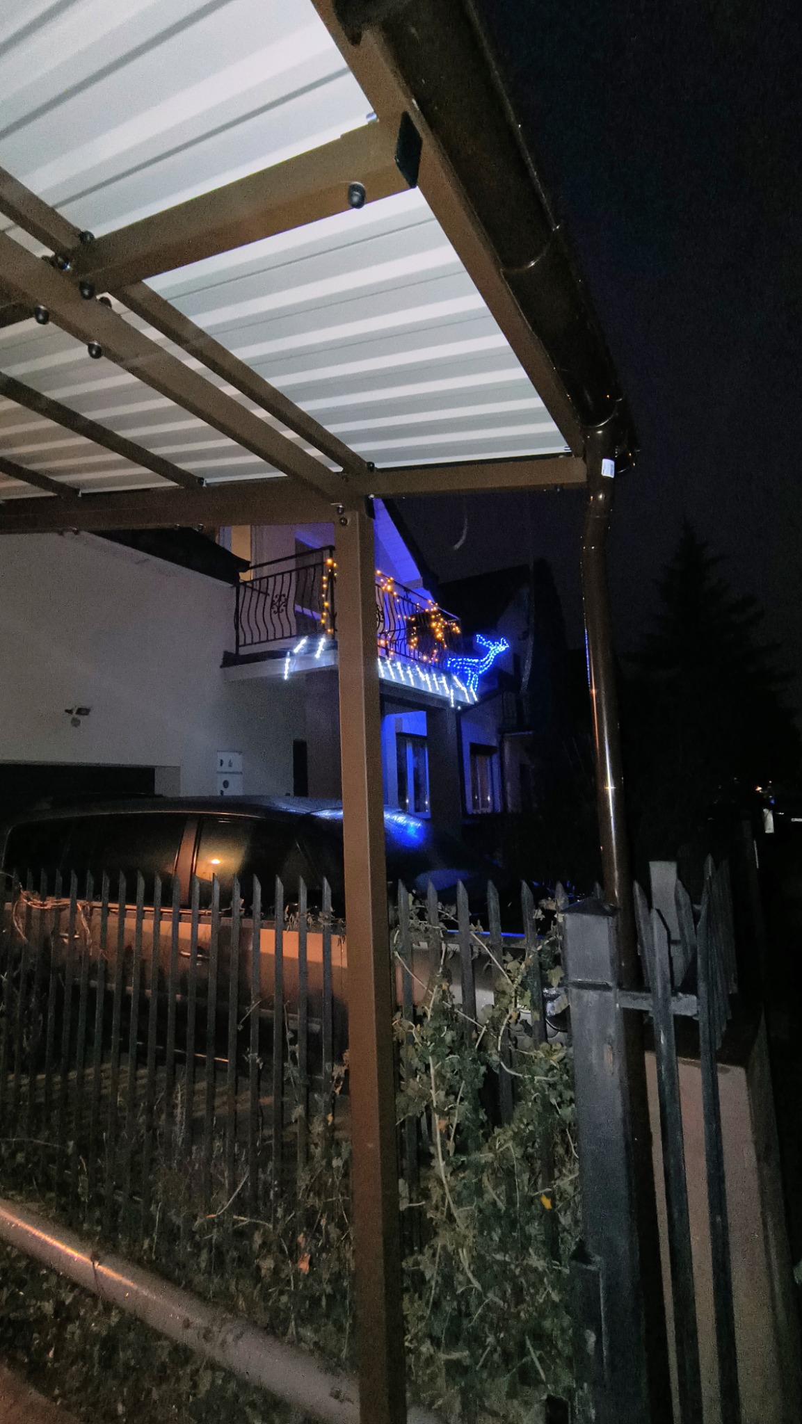 Metalowa konstrukcja z białym zadaszeniem, prawdopodobnie pergola, oświetlona w ciemności. W tle widoczny dom z dekoracjami świątecznymi.