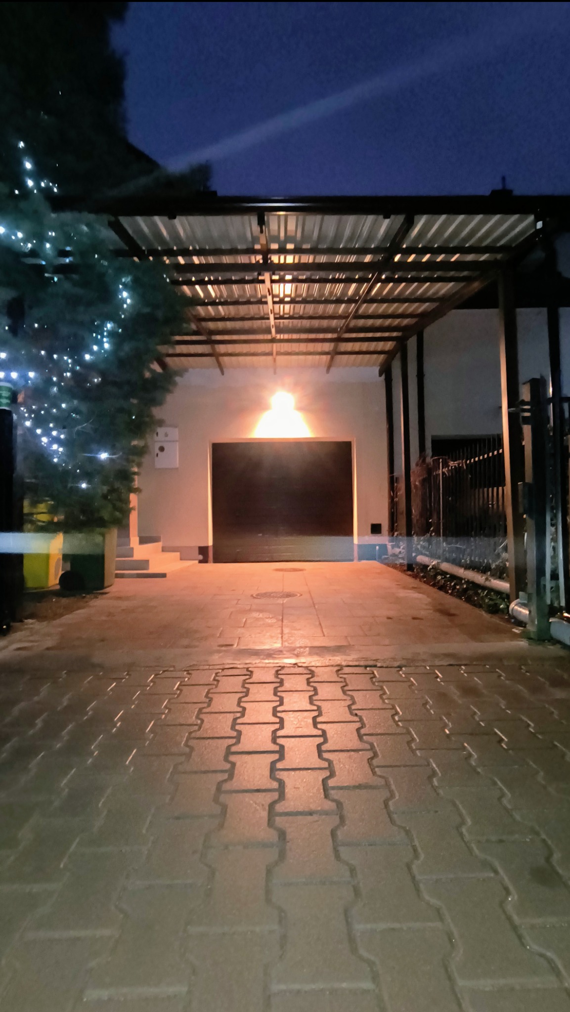 Metalowa pergola zadaszająca podjazd do garażu, oświetlona lampą, obok choinka z lampkami, widok nocą.