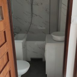 Kamiński Dariusz - Widok na łazienkę z umywalką, toaletą, wanną z prysznicem i pralką, wykończoną płytkami imitującymi marmur, widziany zza częściowo otwartych, drewnianych drzwi.