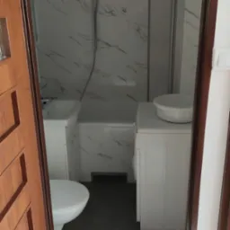 Widok na łazienkę z umywalką, toaletą, wanną z prysznicem i pralką, wykończoną płytkami imitującymi marmur, widziany zza częściowo otwartych, drewnianych drzwi.