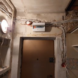 Kamiński Dariusz - Wejście do pomieszczenia w trakcie remontu: widoczne przewody elektryczne na ścianie, niezabezpieczone kable, stara futryna drzwiowa, brak wykończenia ścian.
