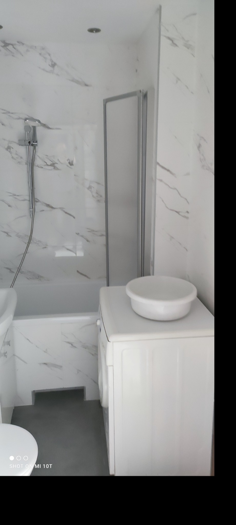 Nowoczesna łazienka z białymi marmurowymi płytkami, kabiną prysznicową ze srebrną ramą, białą pralką z miską na górze i szarą podłogą.