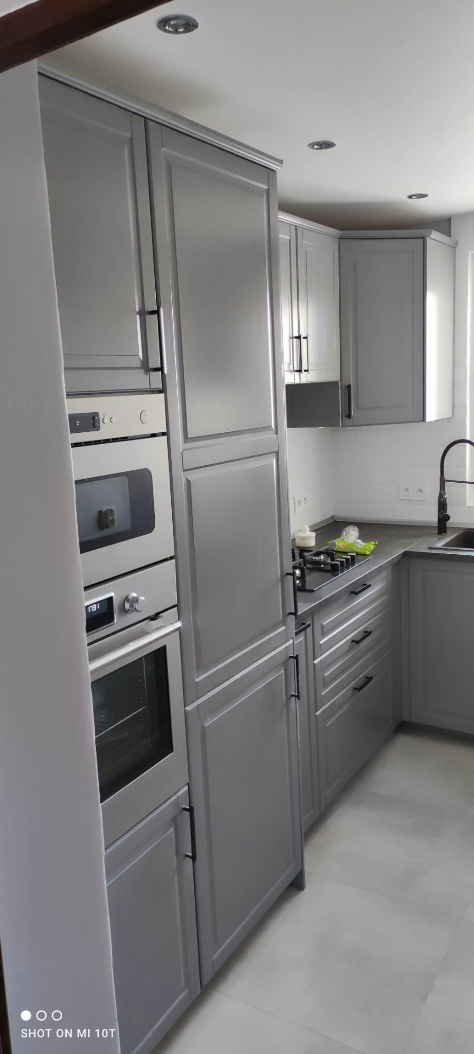 Szara kuchnia z zabudową meblową, piekarnikiem i płytą gazową, czarny kran przy zlewie, płytki na ścianie.