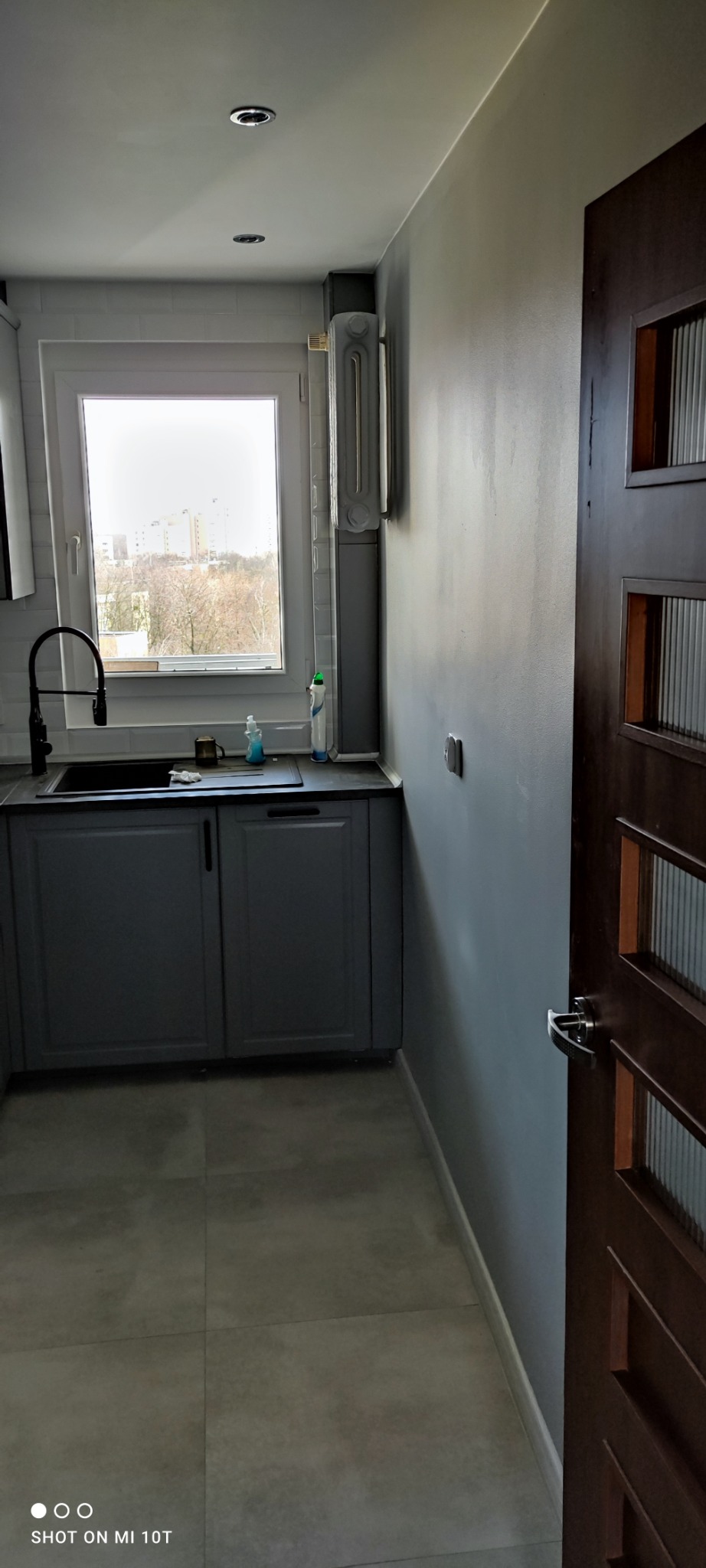 Świeżo wykończona kuchnia w odcieniach szarości, z widokiem na miasto za oknem, drewniane drzwi z przeszkleniami po prawej stronie kadru.