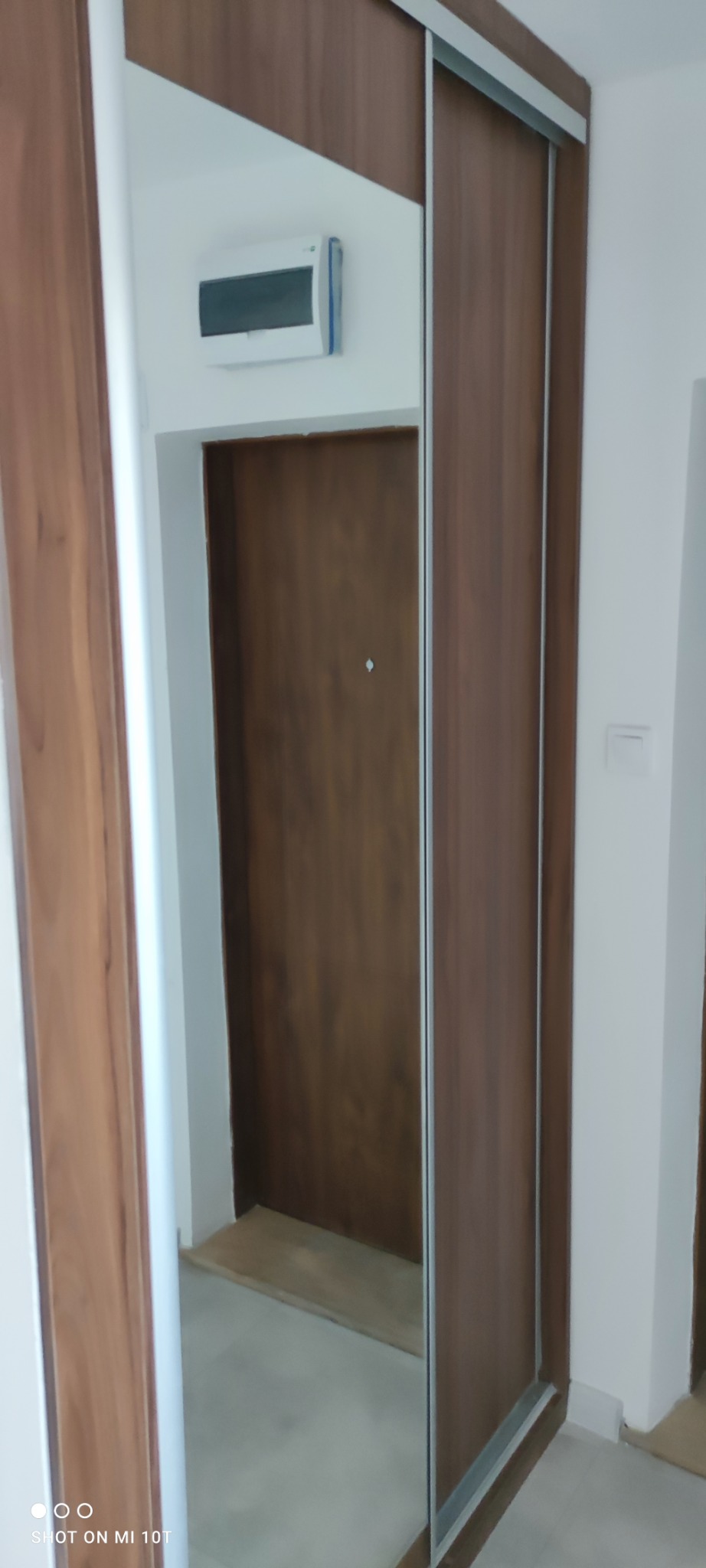 Wnętrze szafy wnękowej z przesuwnymi drzwiami o drewnopodobnym wykończeniu, widoczna również część ściany z domofonem i fragment drzwi wejściowych odbitych w lustrze.