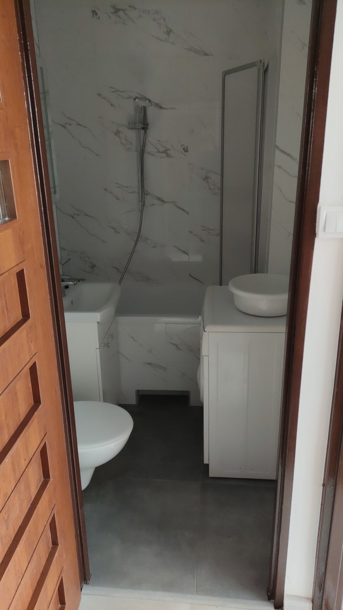 Widok na łazienkę z umywalką, toaletą, wanną z prysznicem i pralką, wykończoną płytkami imitującymi marmur, widziany zza częściowo otwartych, drewnianych drzwi.