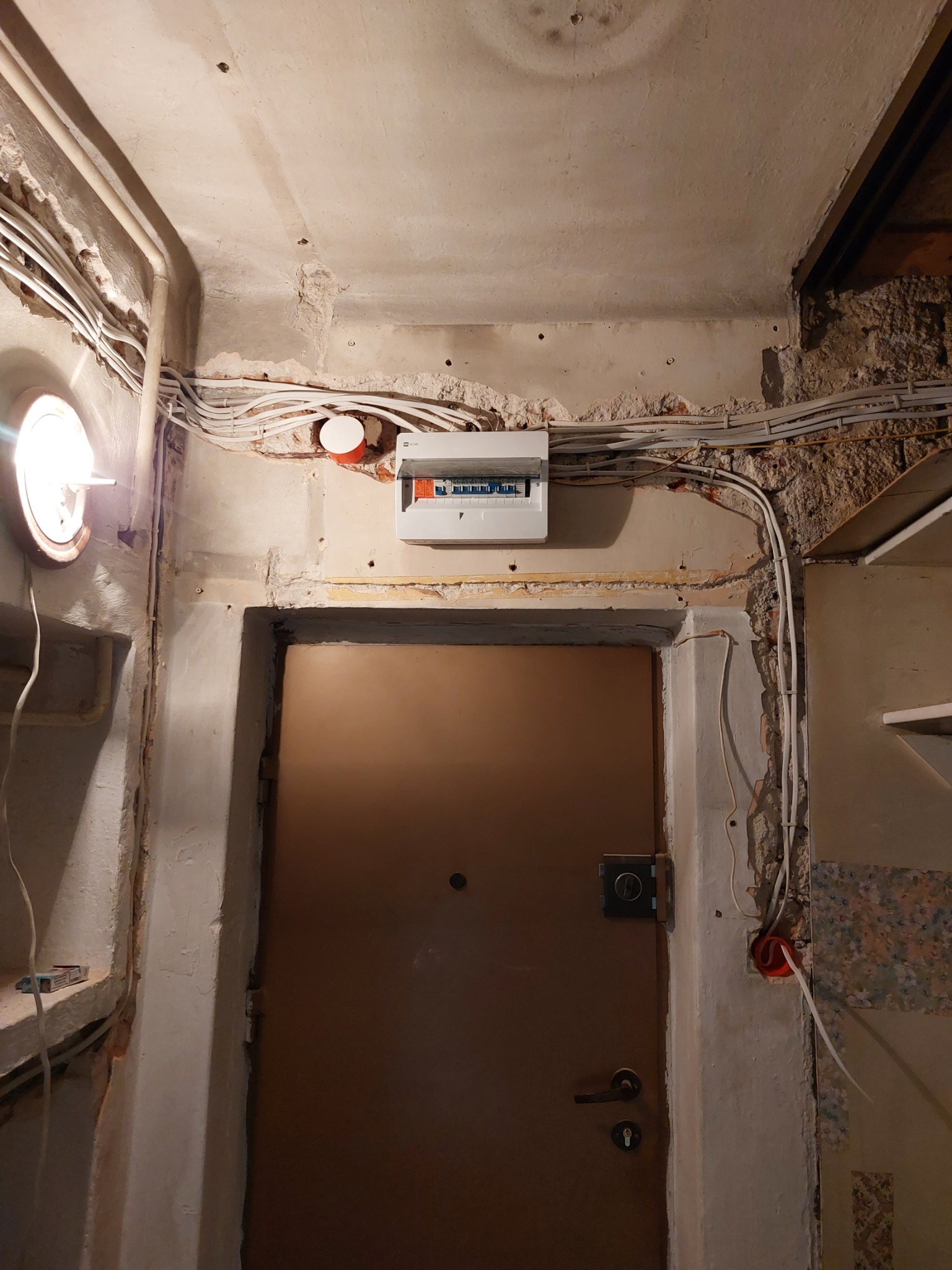 Wejście do pomieszczenia w trakcie remontu: widoczne przewody elektryczne na ścianie, niezabezpieczone kable, stara futryna drzwiowa, brak wykończenia ścian.