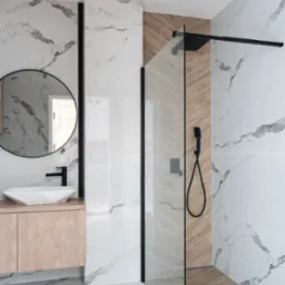 Nowoczesna łazienka w Piątkowisku z marmurowymi płytkami, kabiną prysznicową ze szkła i czarnymi elementami armatury, drewniana szafka pod umywalką z okrągłym lustrem.