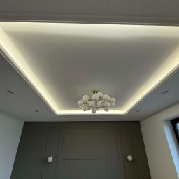 Marciniak Remonty Damian Marciniak - Nowoczesny sufit podwieszany z oświetleniem LED i dekoracyjną lampą w kształcie kuli, szara ściana z ozdobnymi panelami i kinkietami. Wykończenie wnętrza.