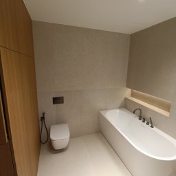 Marciniak Remonty Damian Marciniak - Minimalistyczna łazienka w beżach: wolnostojąca wanna, podwieszana toaleta z bidetką, drewniana zabudowa. Oświetlenie punktowe i wnękowe dodaje ciepła.