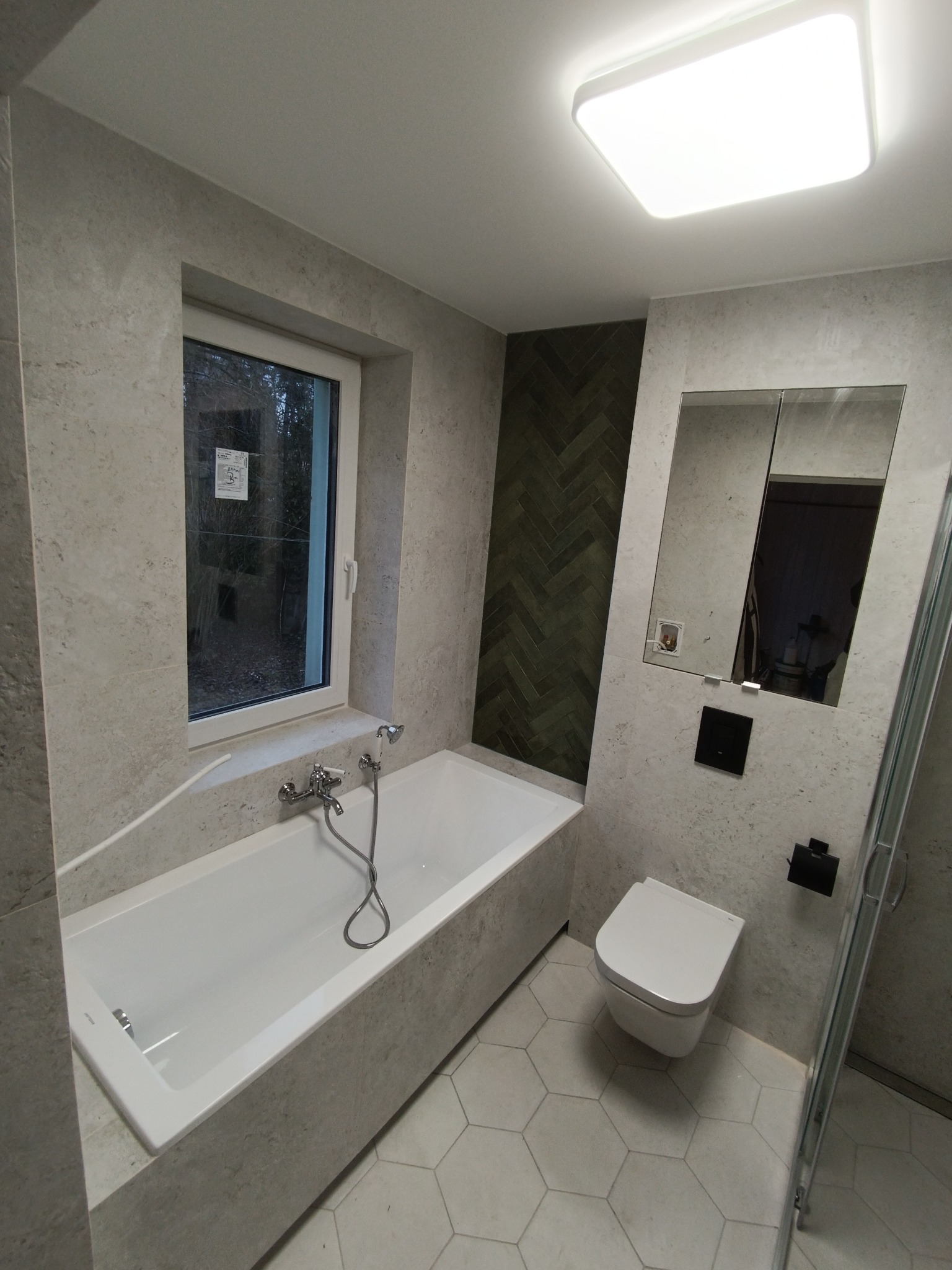 Nowoczesna łazienka z wanną obudowaną kamieniem, szarą mozaiką w jodełkę na ścianie, wiszącą toaletą i podłogą z heksagonalnych płytek.