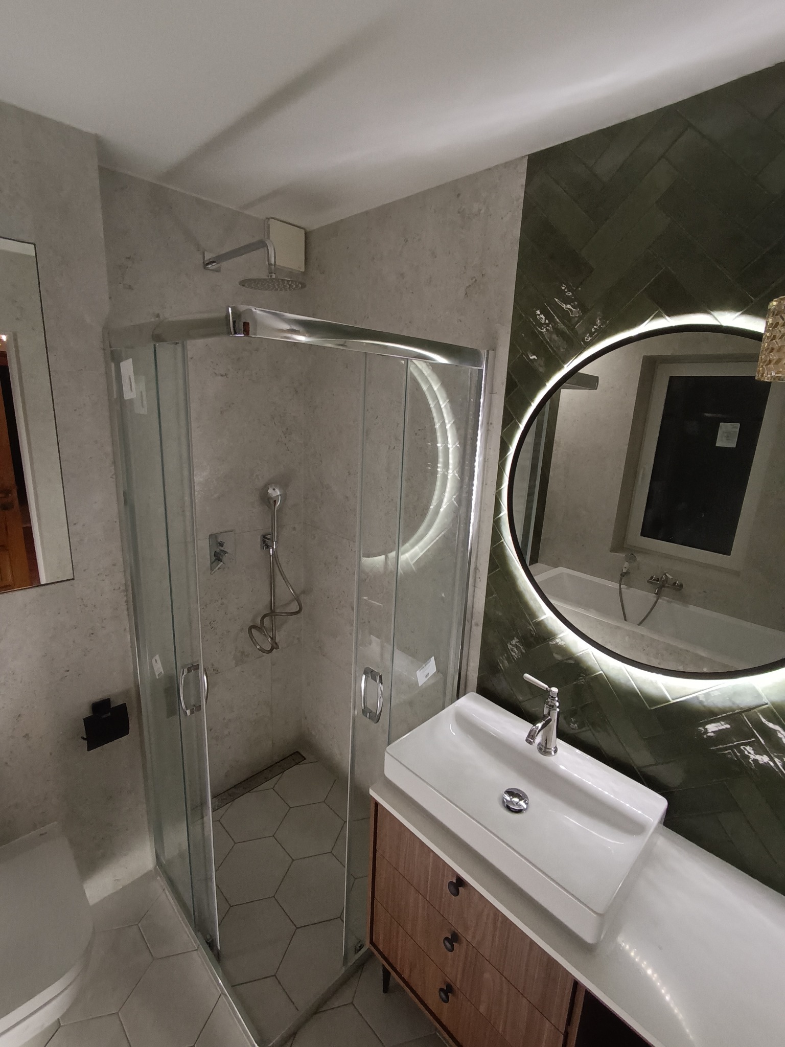 Nowoczesna łazienka z kabiną prysznicową, umywalką nablatową na drewnianej szafce i okrągłym lustrem z podświetleniem LED na ścianie wyłożonej zielonymi kafelkami w jodełkę.