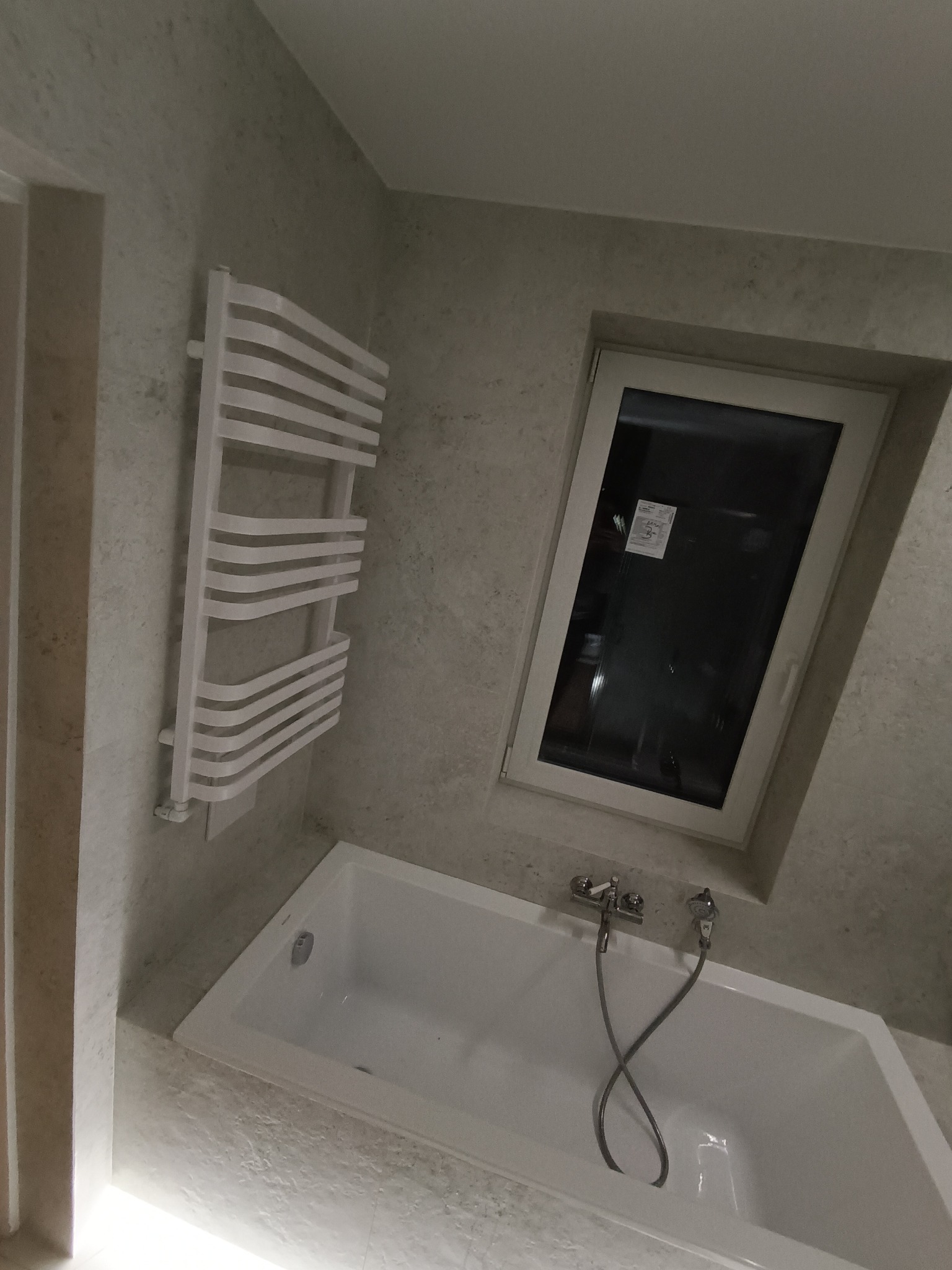 Nowoczesna łazienka z białą wanną, grzejnikiem drabinkowym i oknem, wykończona szarą, wzorzystą płytką imitującą kamień.