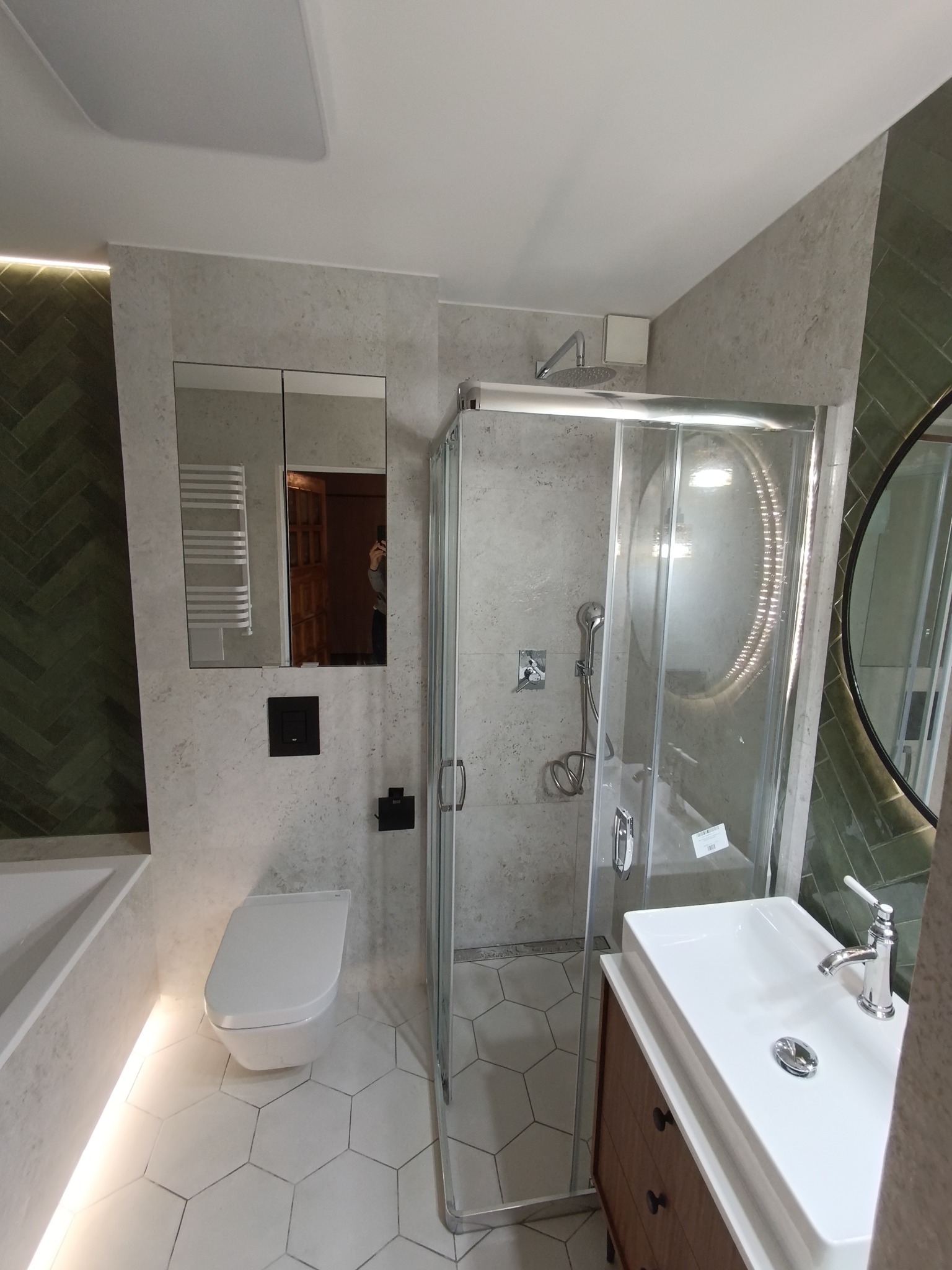 Nowoczesna łazienka z kabiną prysznicową, umywalką na szafce z drewnianymi szufladami, wiszącą toaletą i lustrem z szafką wbudowanym w szarą ścianę z imitacją kamienia, zielona ściana w jodełkę...