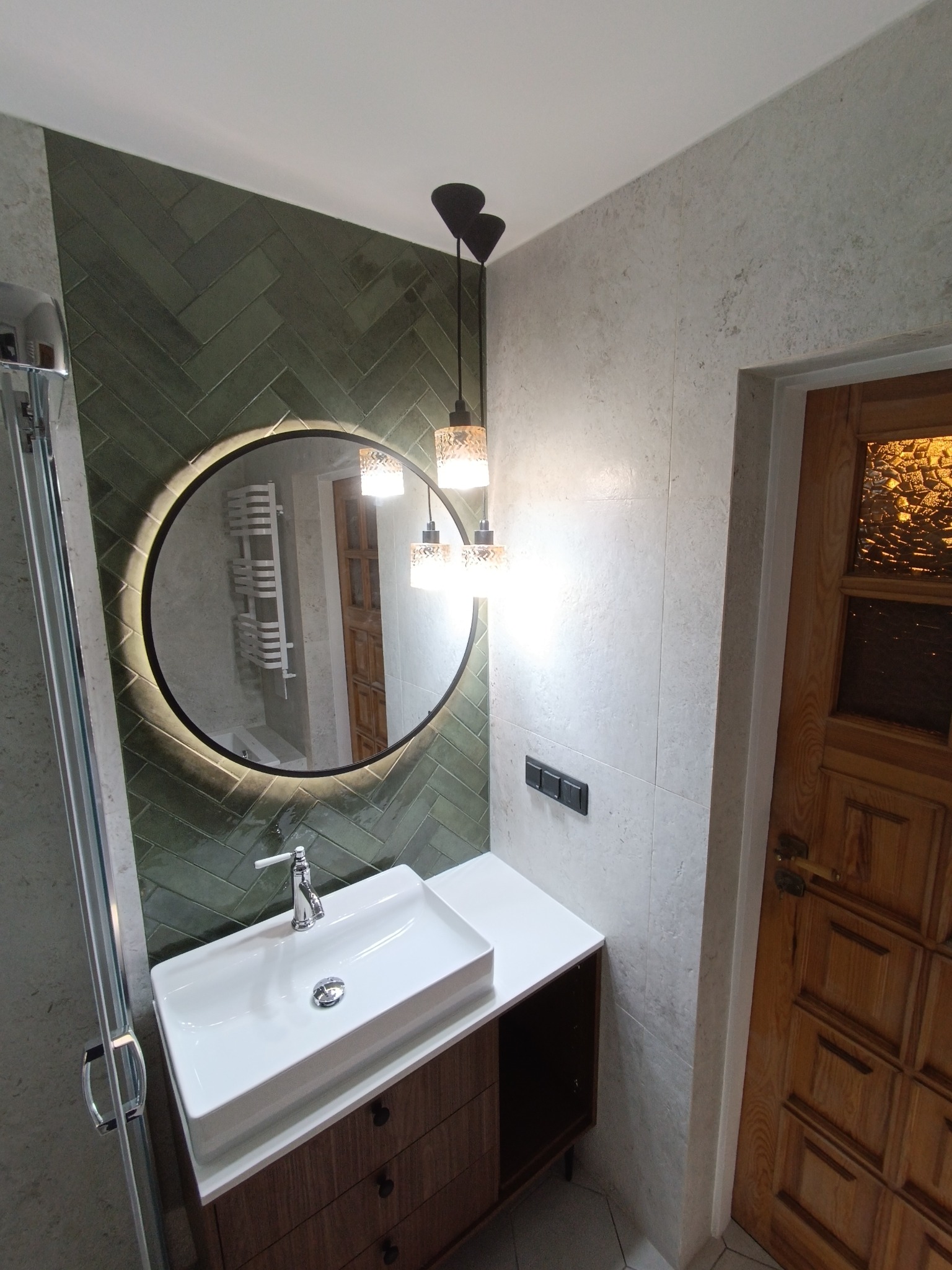 Nowoczesna łazienka z umywalką nablatową na szafce z drewnianymi szufladami, okrągłe lustro z podświetleniem LED, zielone płytki ułożone w jodełkę na ścianie, wiszące lampy z ozdobnymi kloszami...