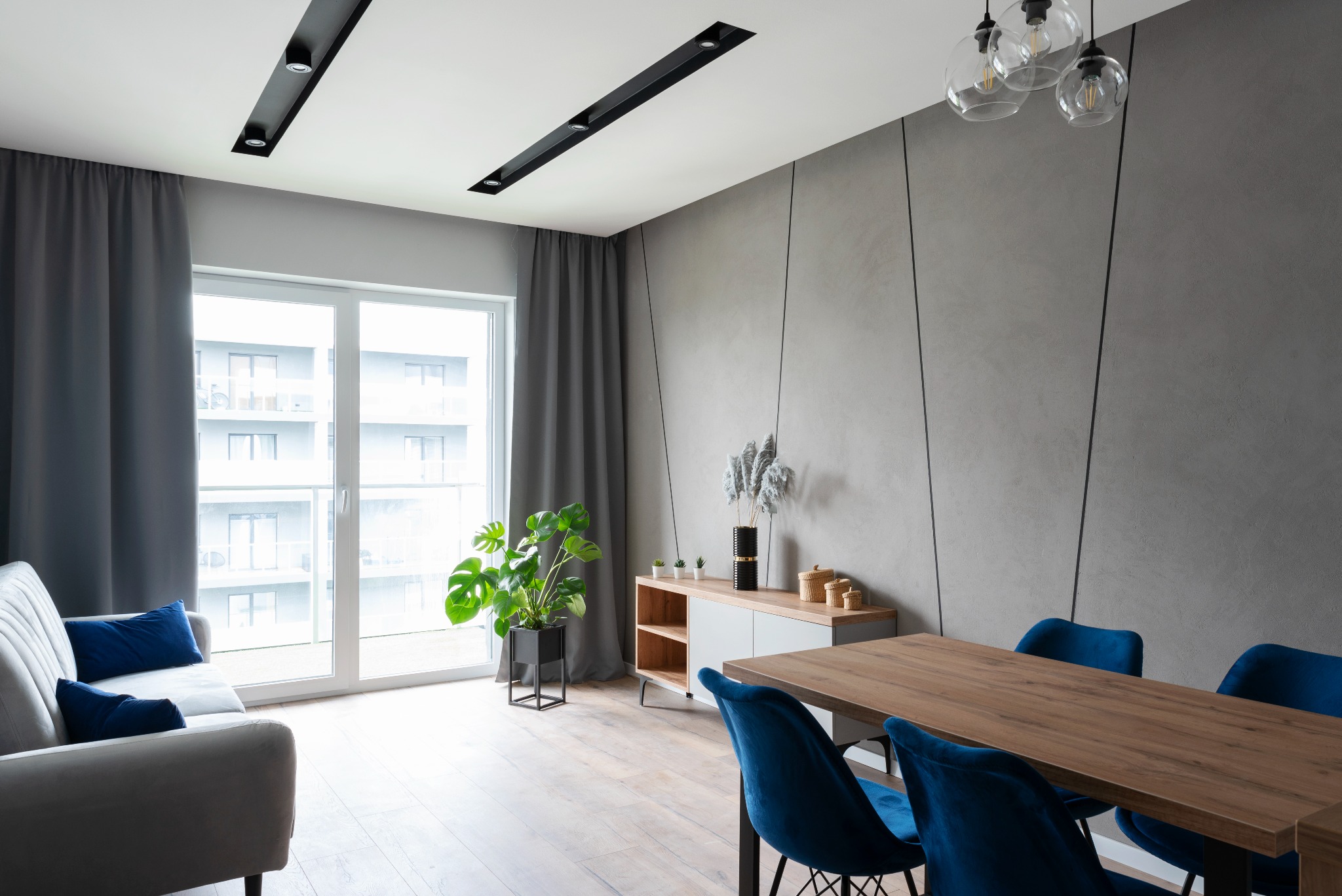 Nowoczesny salon z szarą ścianą z dekoracyjnymi liniami, drewnianym stołem z aksamitnymi krzesłami, jasną sofą z niebieskimi poduszkami i dużym oknem balkonowym z szarymi zasłonami.