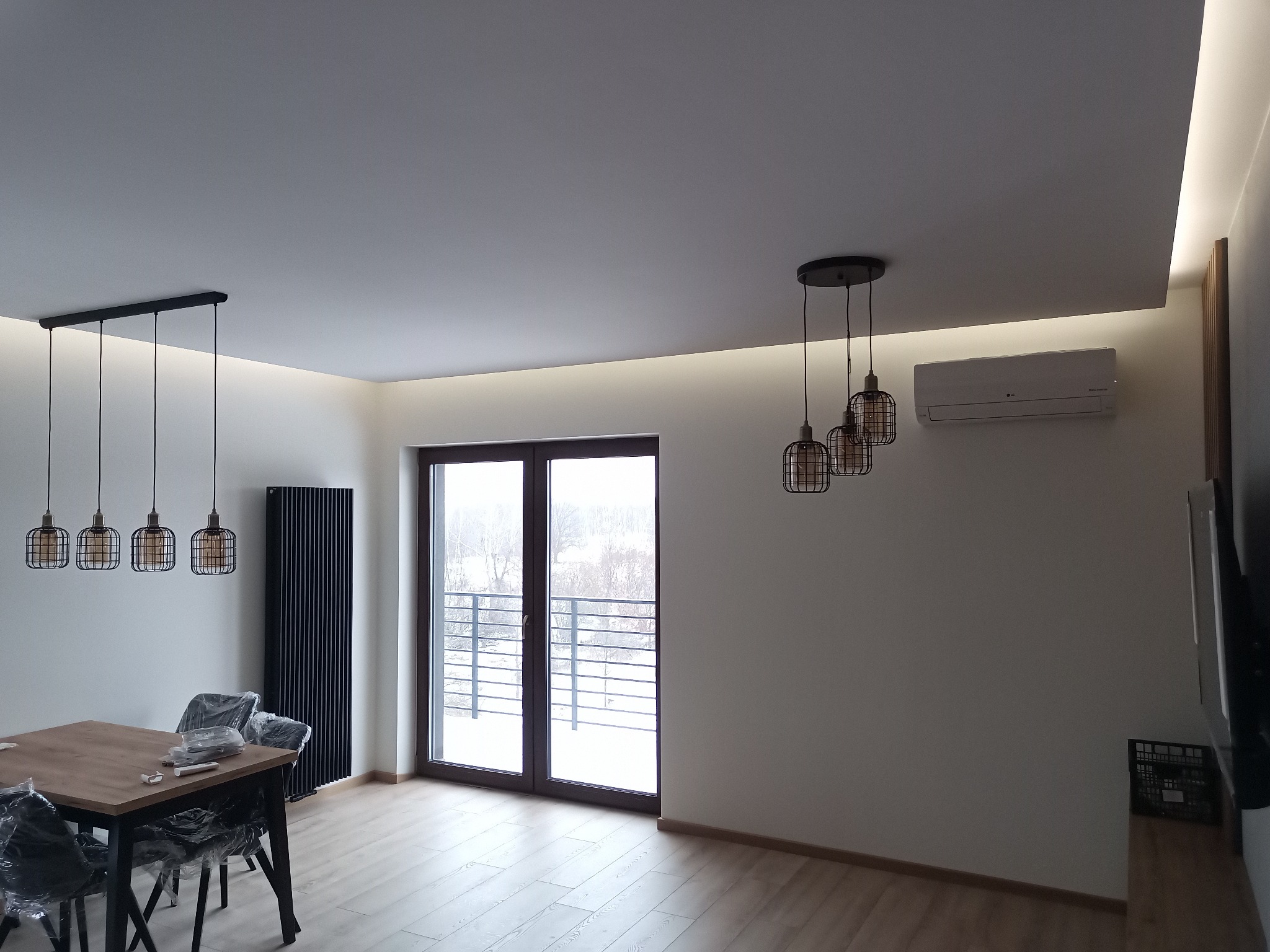 Nowoczesne wnętrze pokoju dziennego z drewnianym stołem, czarnymi krzesłami w folii, wiszącymi lampami w stylu industrialnym, grzejnikiem panelowym i oknem balkonowym z widokiem na zimowy krajobraz.