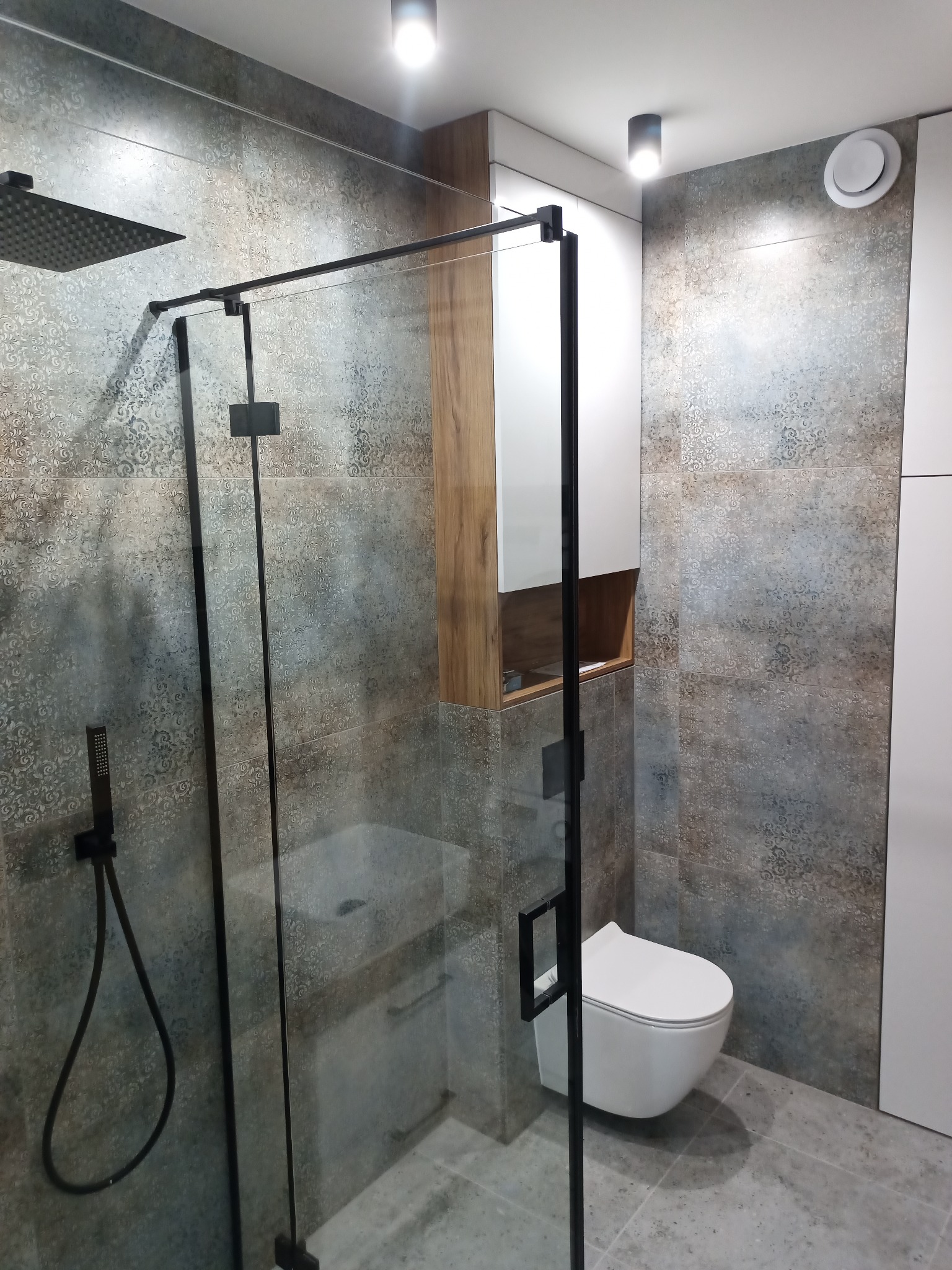Nowoczesna łazienka z kabiną prysznicową typu walk-in z czarnymi profilami, wiszącą toaletą i szafką z drewnianymi elementami, wykończona płytkami w odcieniach szarości z delikatnym wzorem.