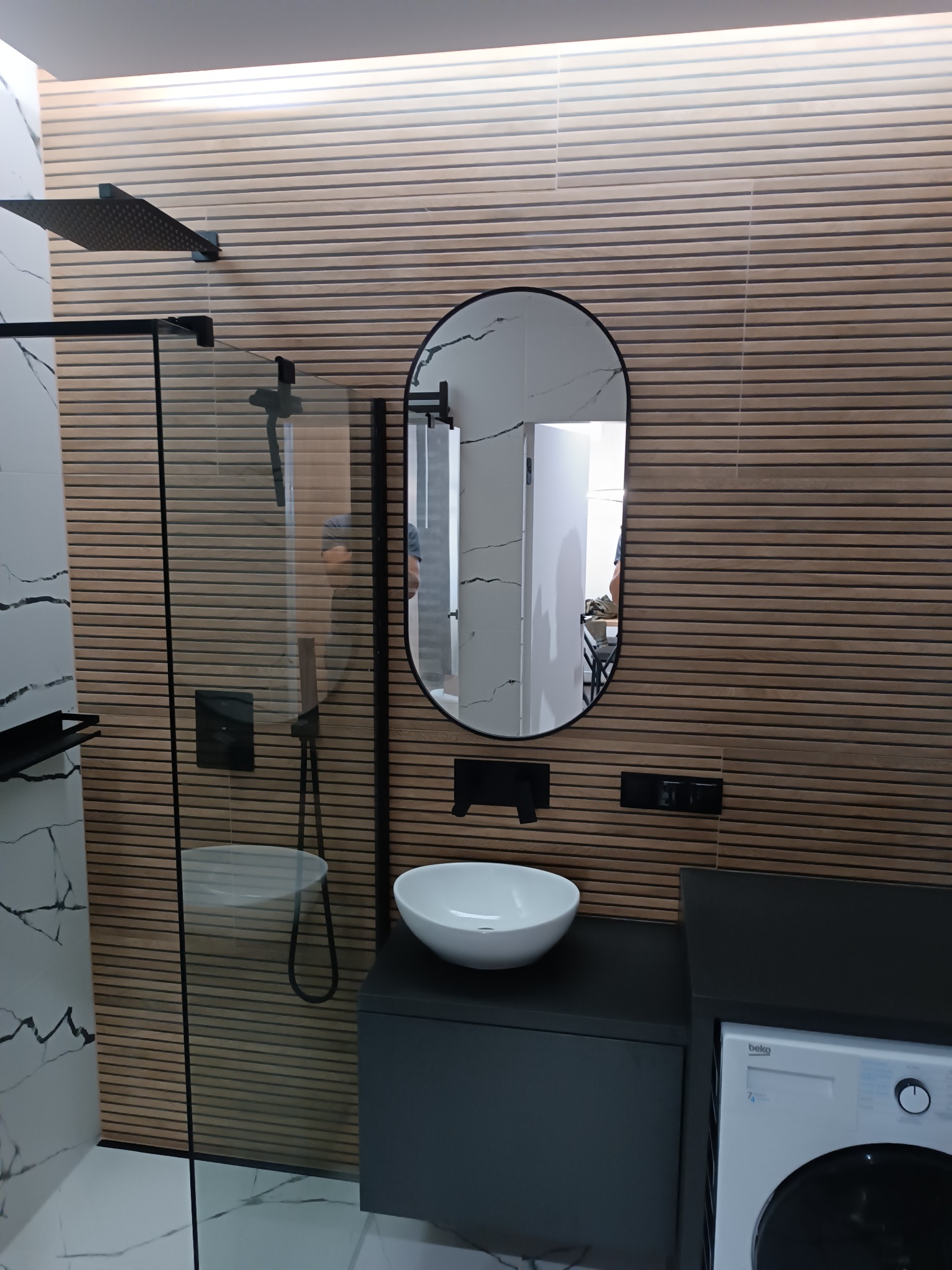 Nowoczesna łazienka z kabiną prysznicową typu walk-in, umywalką nablatową na szafce w kolorze grafitowym oraz pralką. Ściana wykończona panelami drewnopodobnymi z poziomymi żłobieniami. W lustrze...