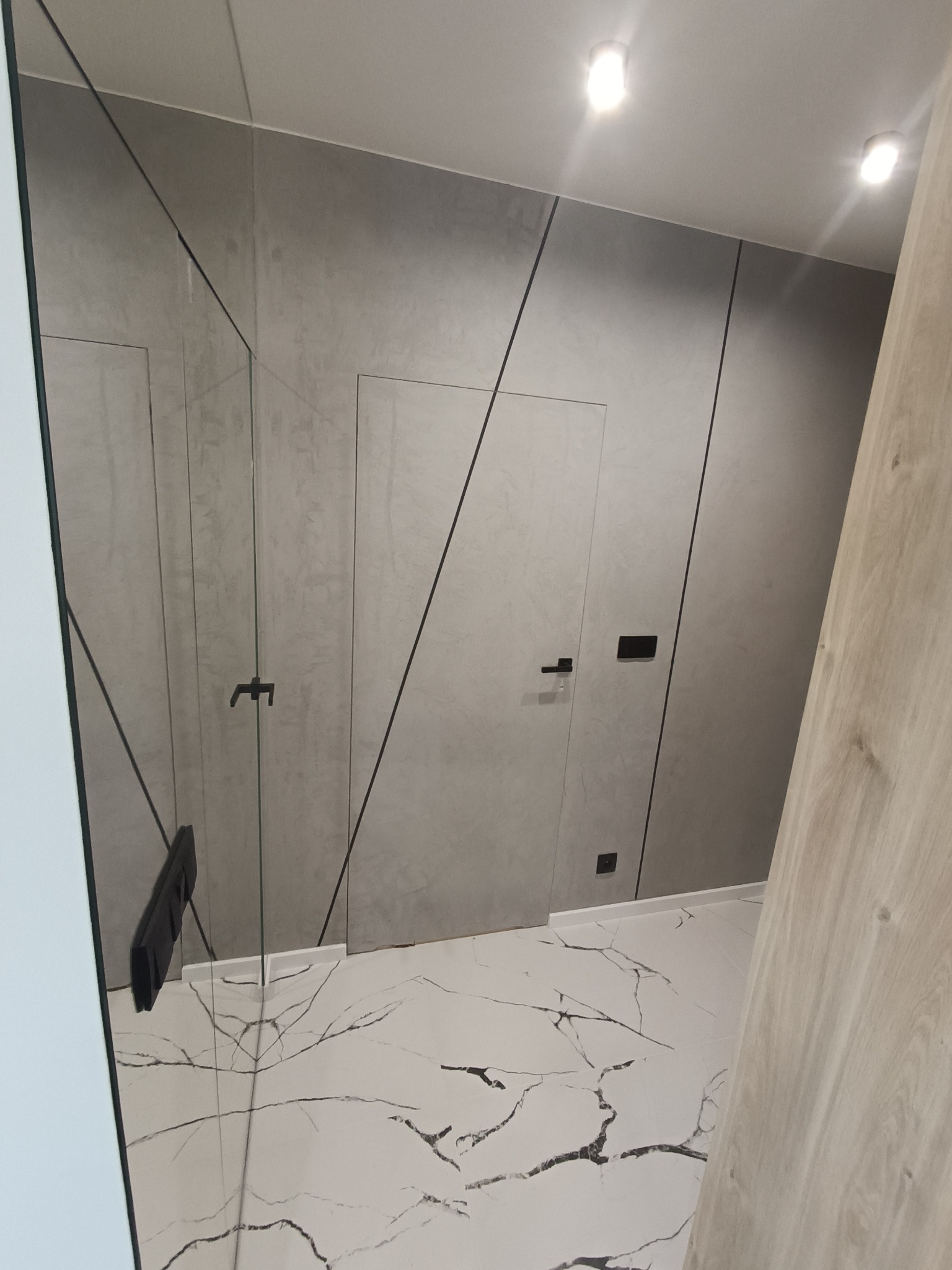 Nowoczesna łazienka z szarą ścianą o fakturze betonu, ukrytymi drzwiami i podłogą z białego marmuru z czarnymi żyłami, oświetlona punktowymi lampami sufitowymi.