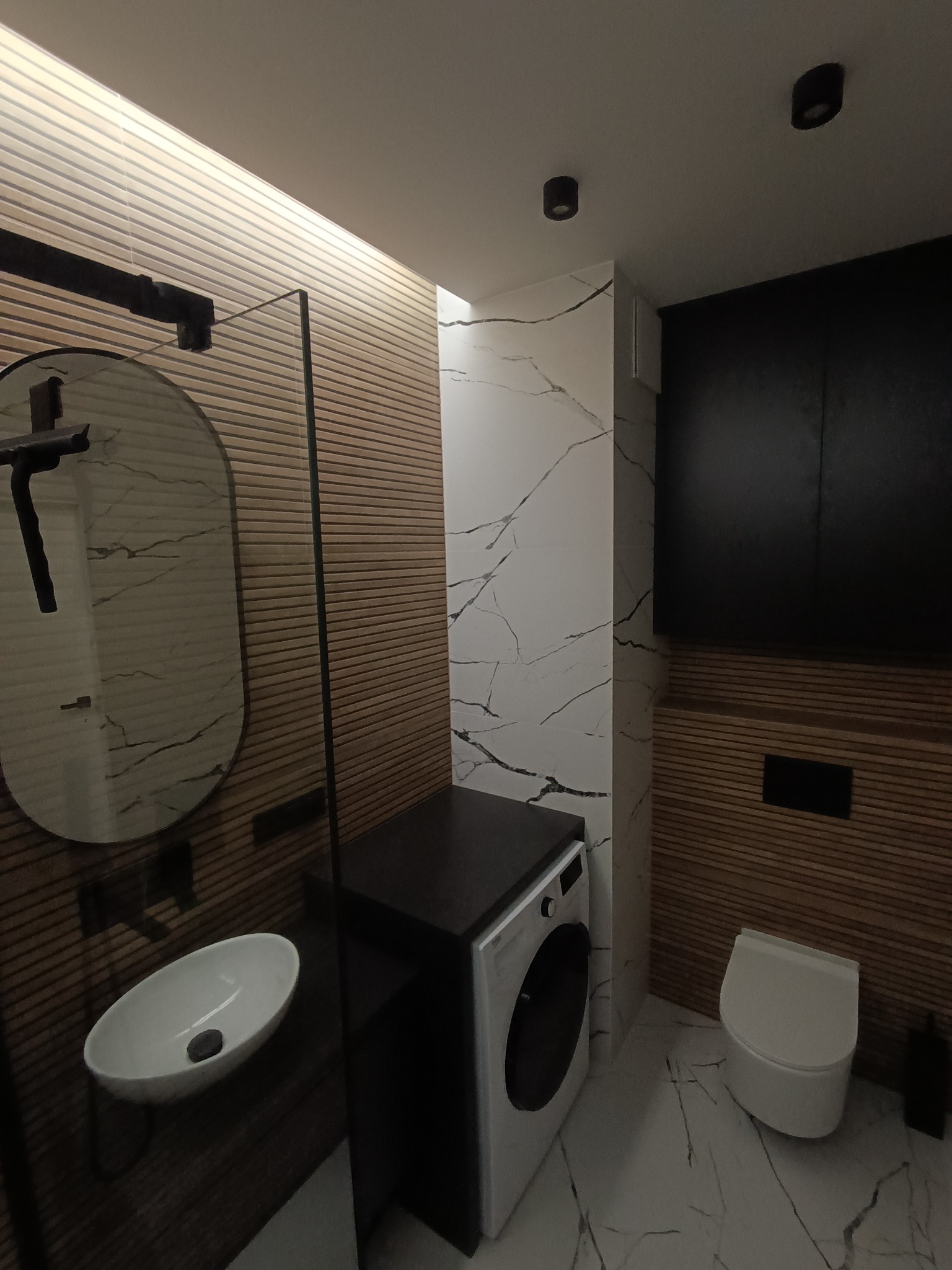 Nowoczesna łazienka z umywalką nablatową, okrągłym lustrem z czarną ramą, pralką pod blatem i toaletą wiszącą, wykończona płytkami imitującymi drewno i marmur.