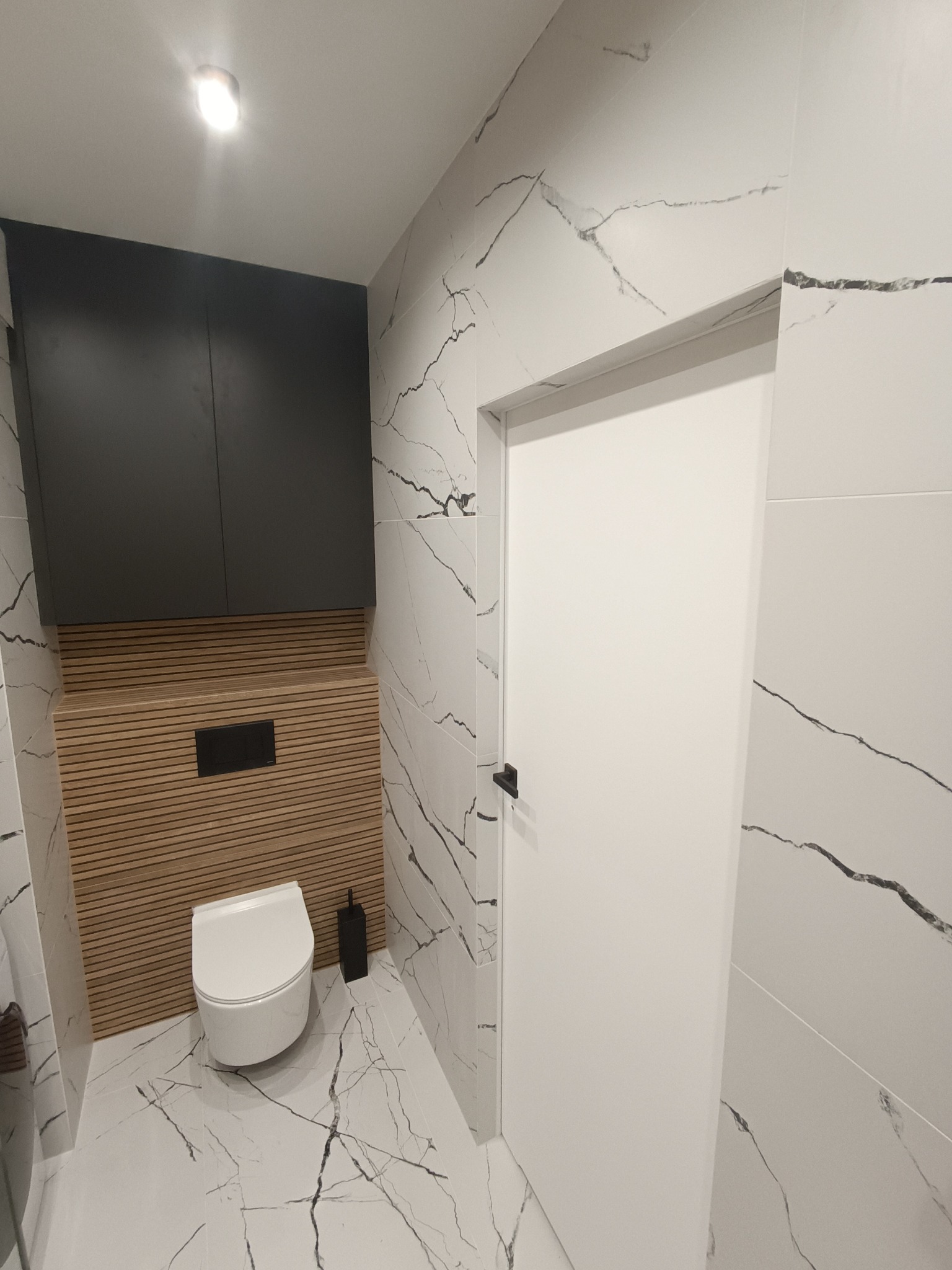 Nowoczesna łazienka z toaletą podwieszaną, zabudową meblową w kolorze grafitowym nad ukrytą spłuczką, wykończona płytkami imitującymi biały marmur z czarnymi żyłami i drewnianą boazerią.